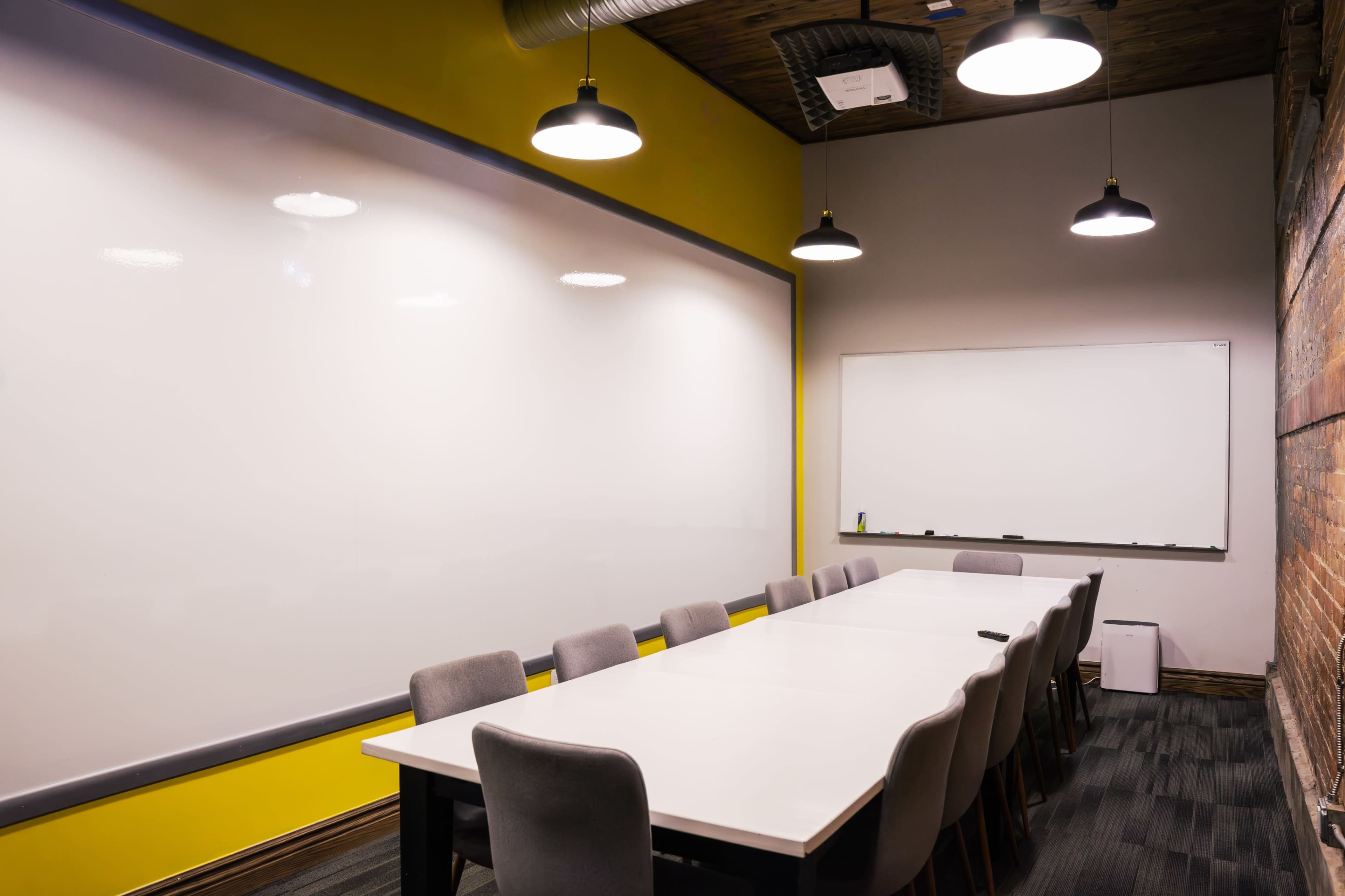 A large conference room features a long white table surrounded by upholstered chairs, with bright yellow walls and a whiteboard at one end.