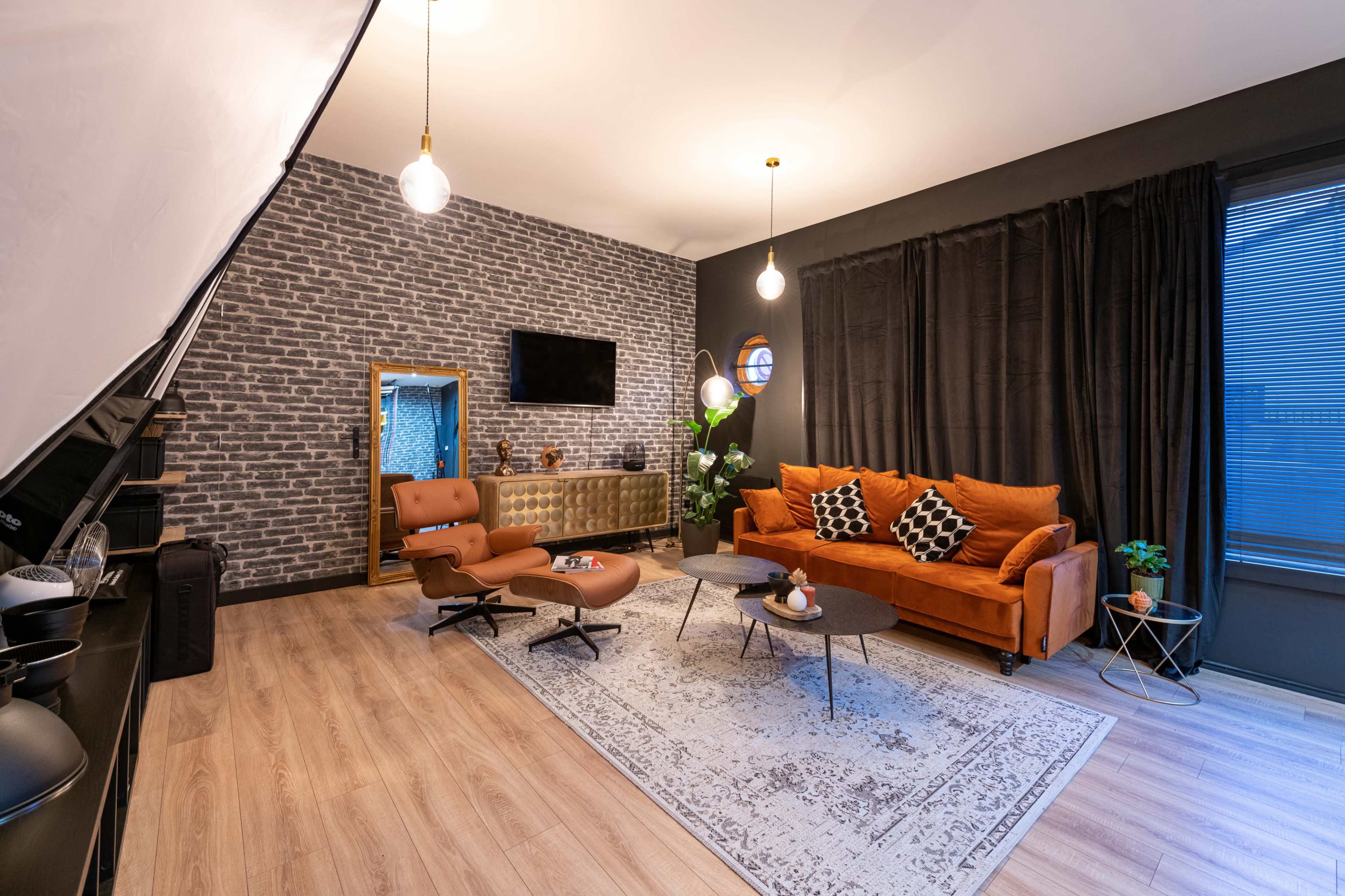 The image shows a modern living room with an orange sofa, a brown lounge chair, a patterned rug, and a brick accent wall.