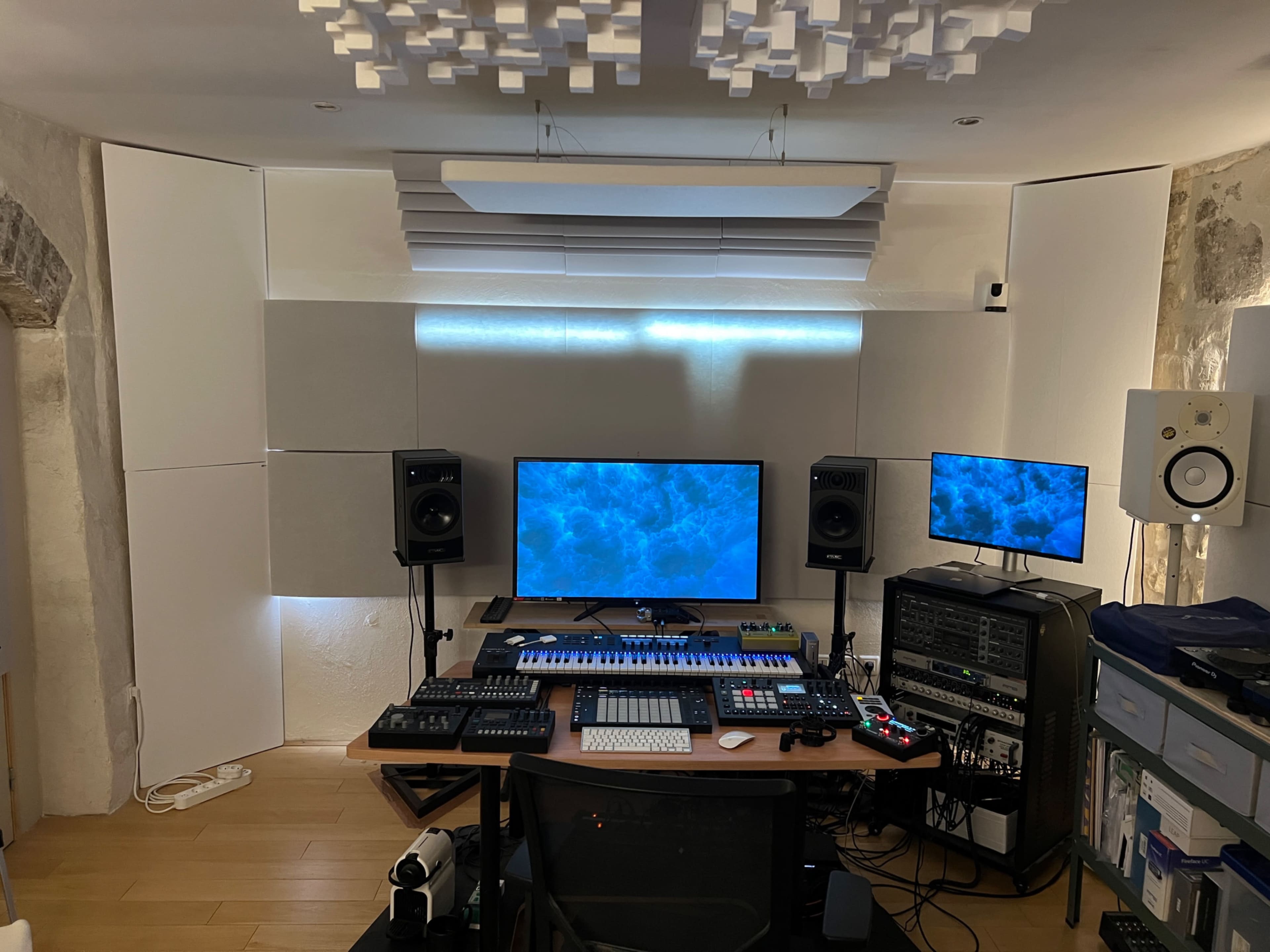 The image shows a music production studio with multiple monitors, audio equipment, and soundproof wall panels arranged around a central desk.