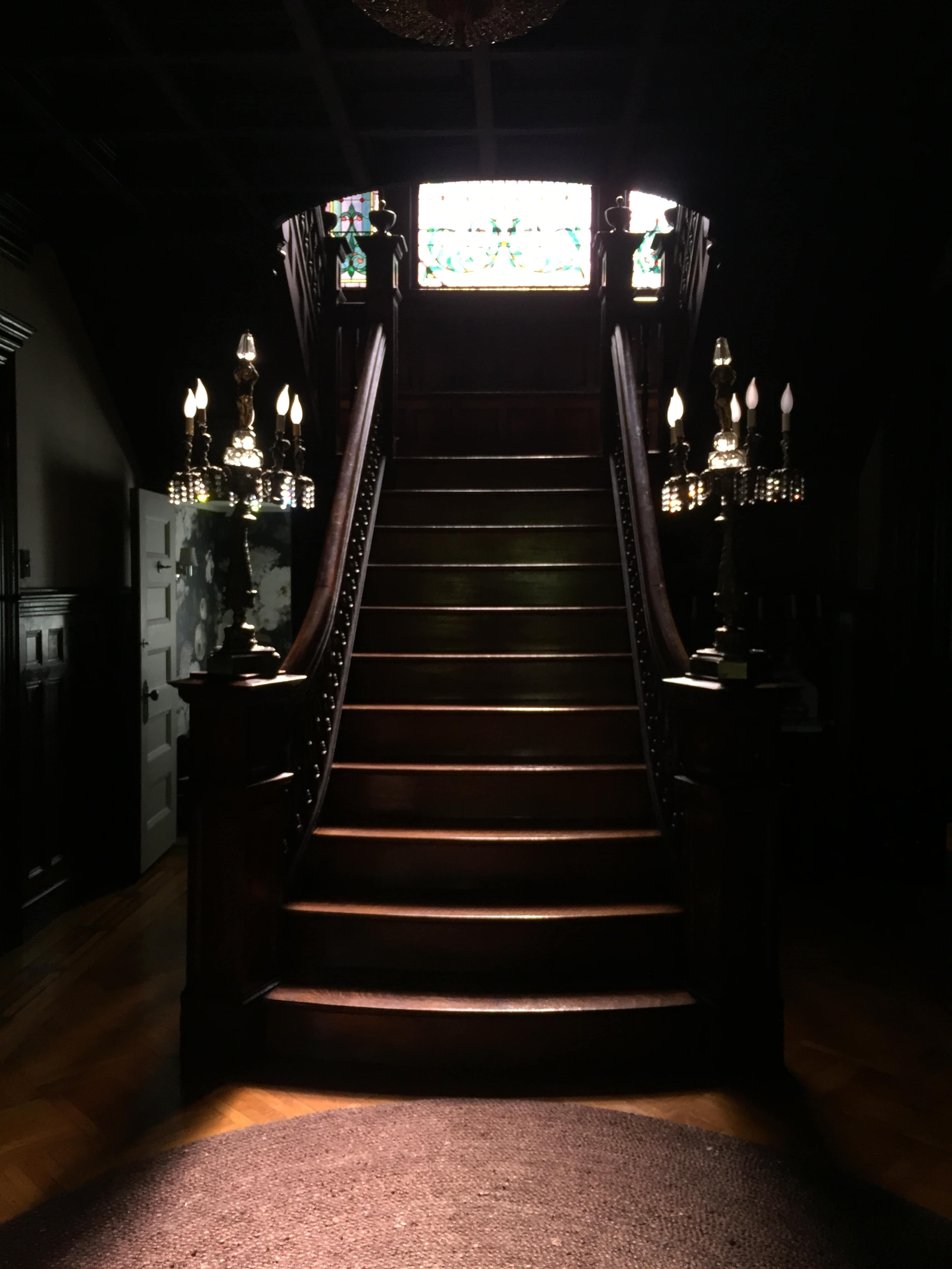 A staircase with ornate wooden railings leads up to a stained glass window illuminated by overhead light.