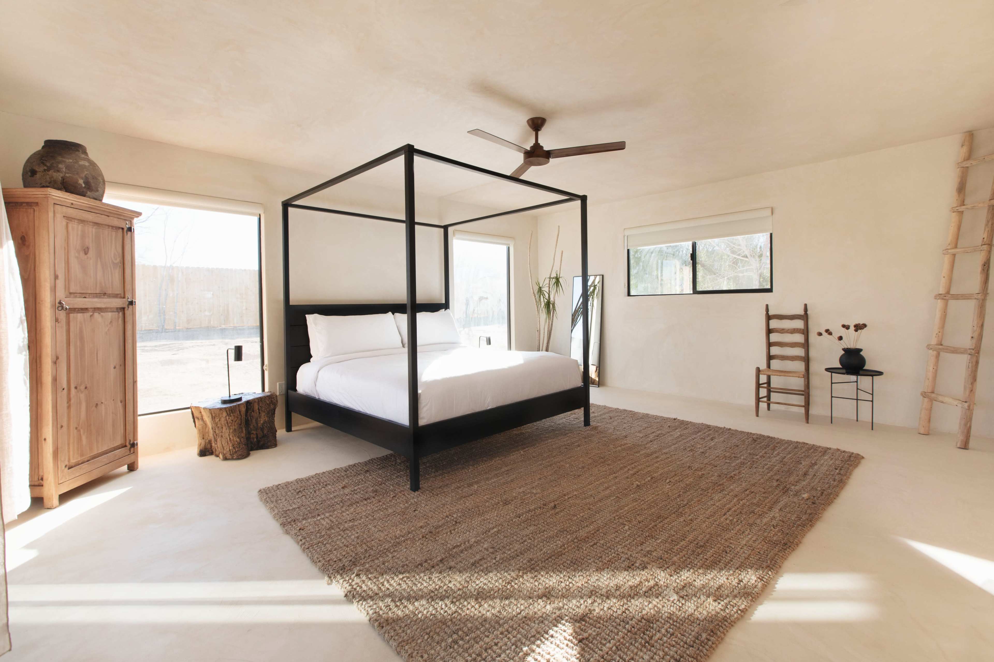 A minimalistic bedroom featuring a black four-poster bed, a woven rug, and large windows allowing natural light to enter.