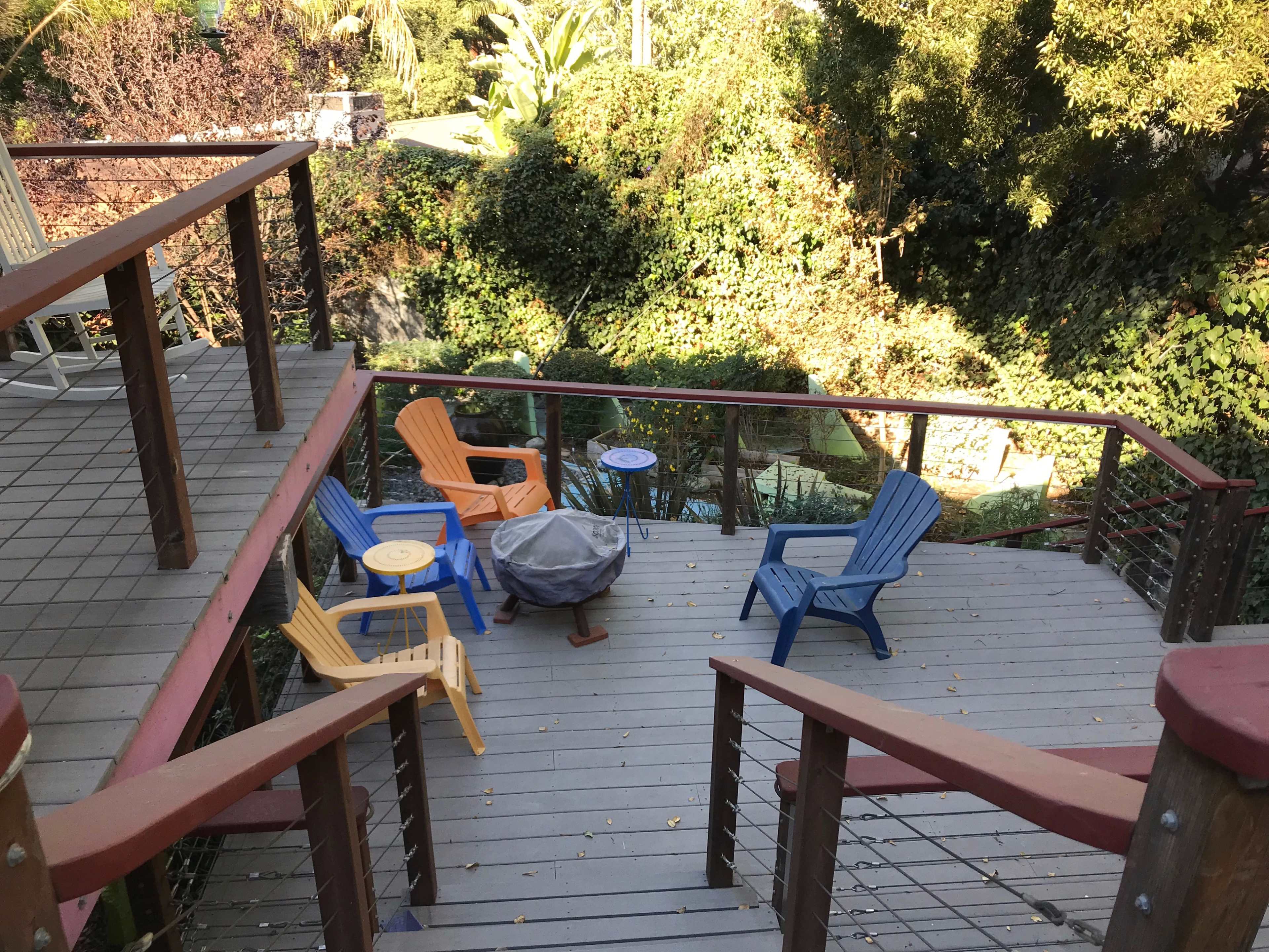 A wooden deck with brightly colored chairs surrounds a small table and fire pit, overlooking a lush, green garden.