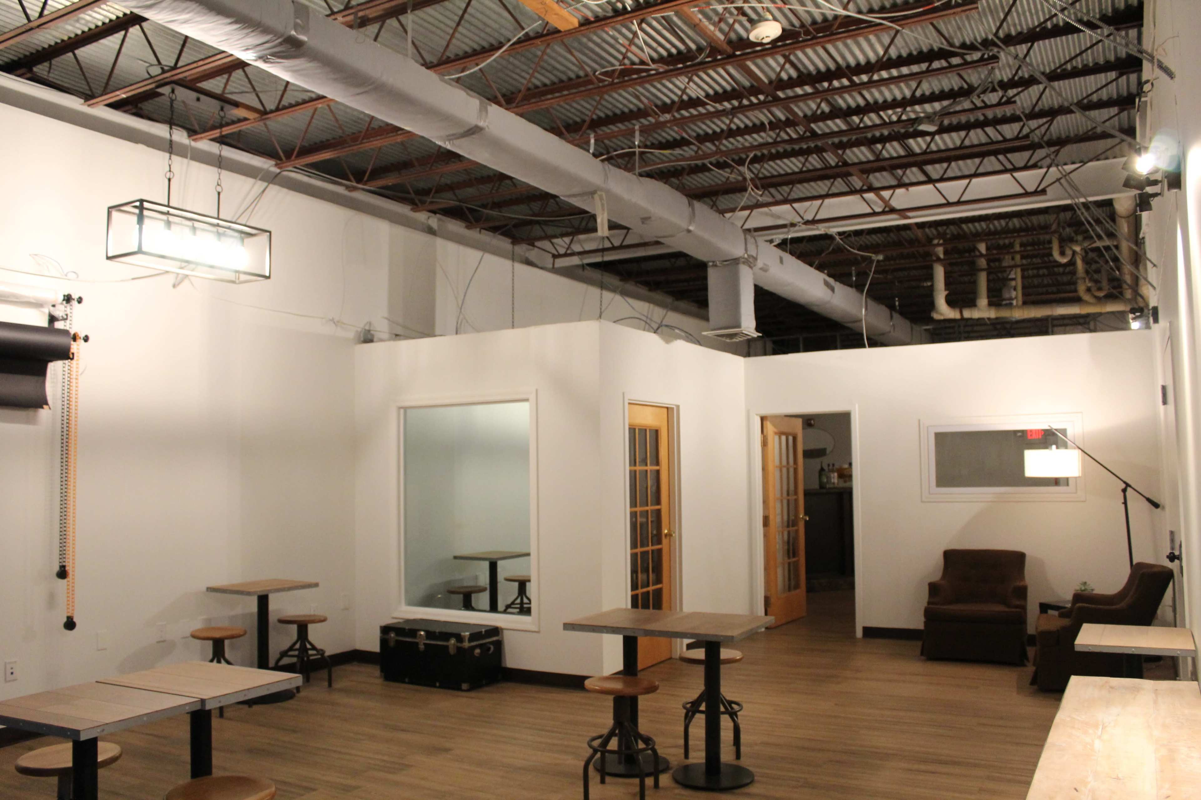An empty industrial-style space with wooden floors, several small tables, and a seating area featuring a chair and side table.