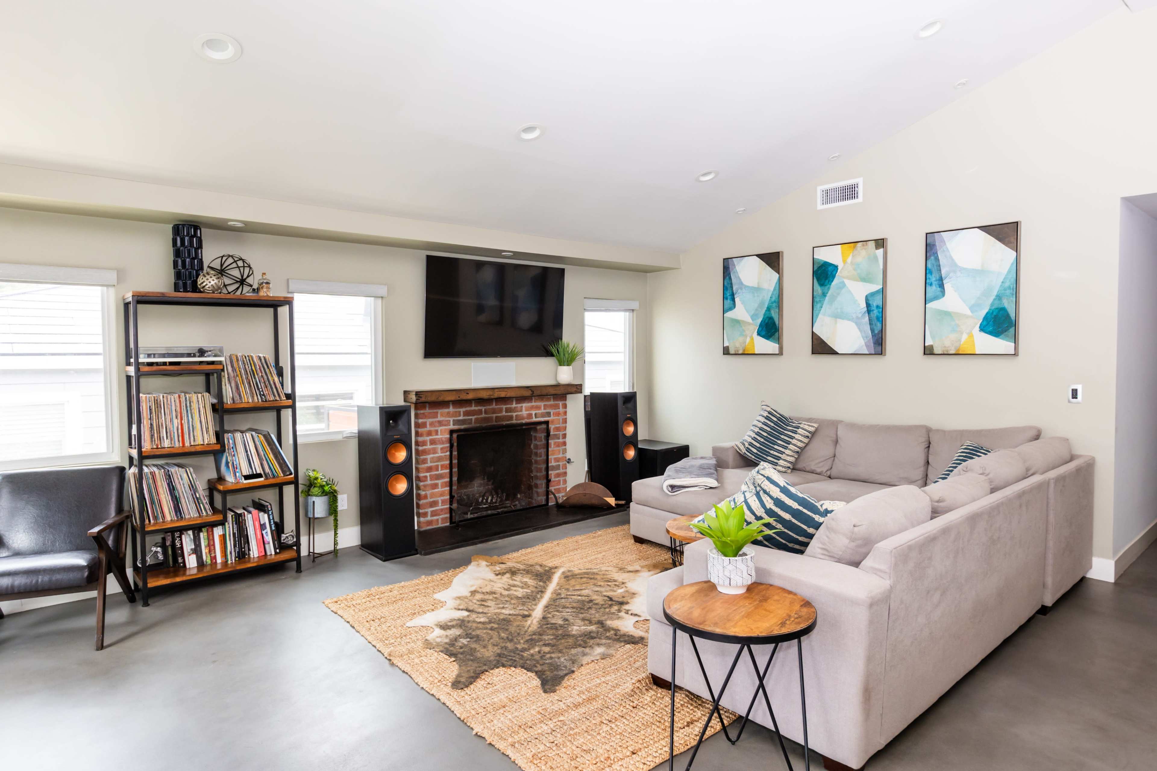 A modern living room features a gray sectional sofa, a wooden bookshelf filled with records, and a brick fireplace with a television mounted above it.
