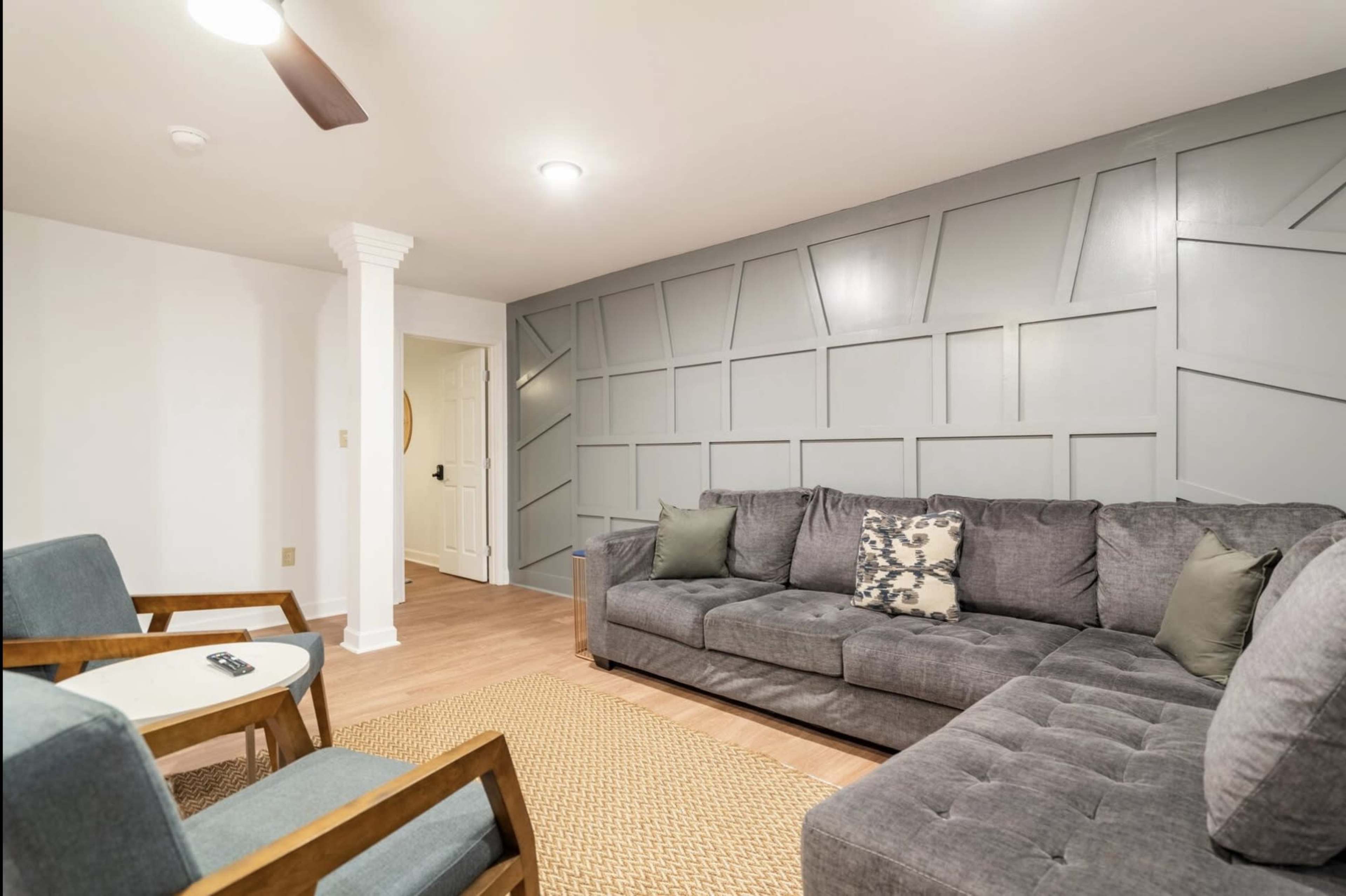 The image shows a living room with a gray sectional sofa, two wooden chairs, and a textured area rug on a light wood floor, against a backdrop of a geometric-patterned gray wall.