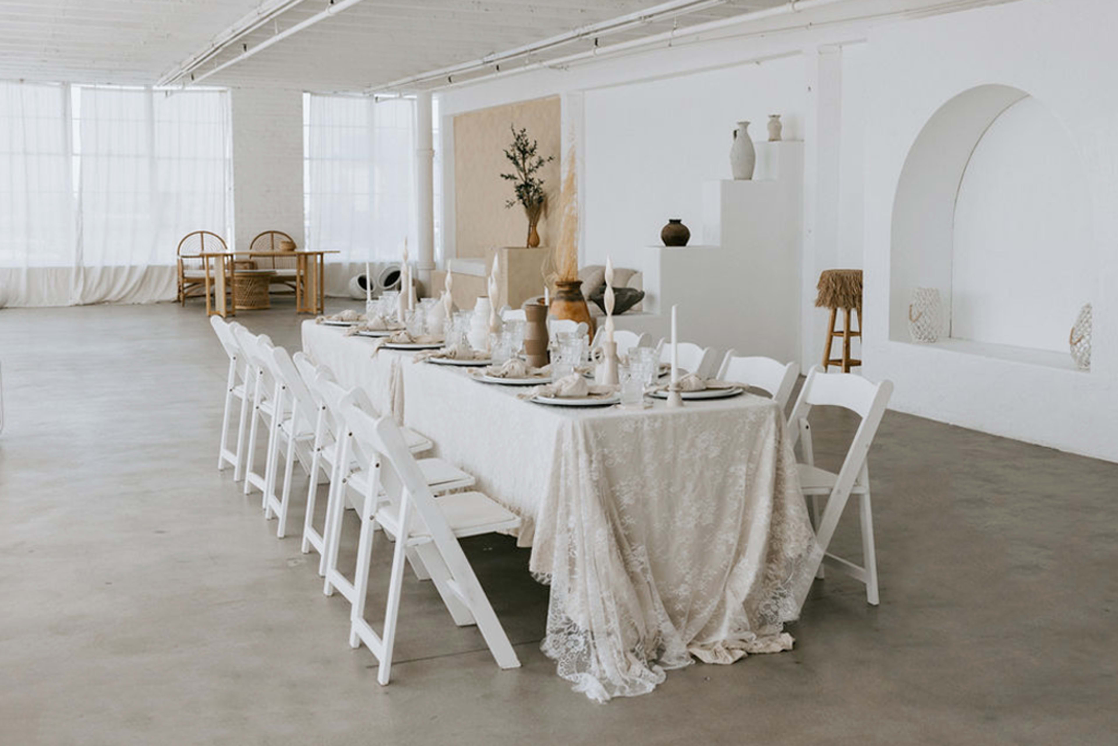 A long dining table is set with white tableware and surrounded by white folding chairs in a bright, minimalistic space.
