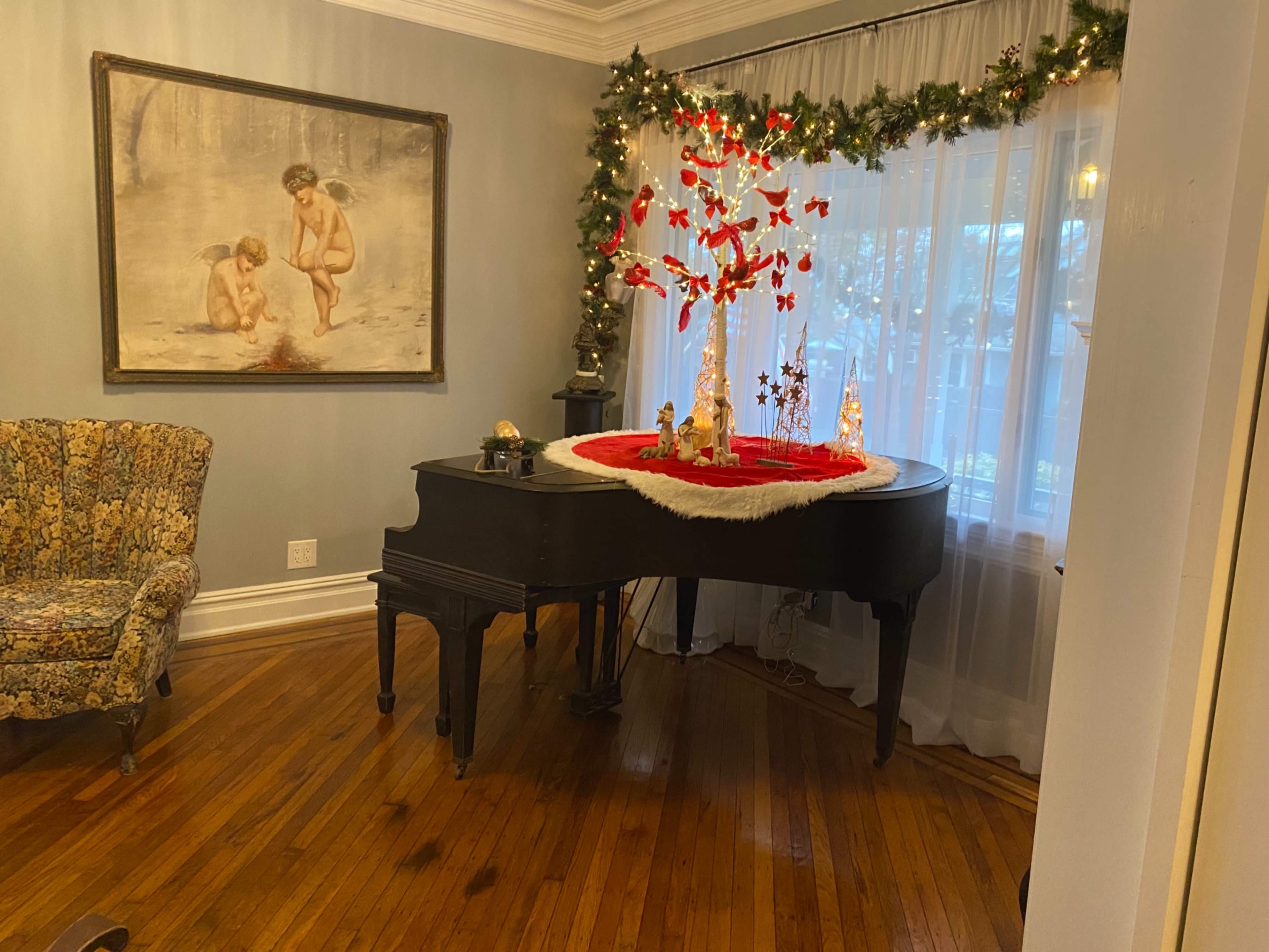A grand piano is centered in a room decorated with a red and white holiday setting, surrounded by a floral-patterned armchair and a large painting on the wall.