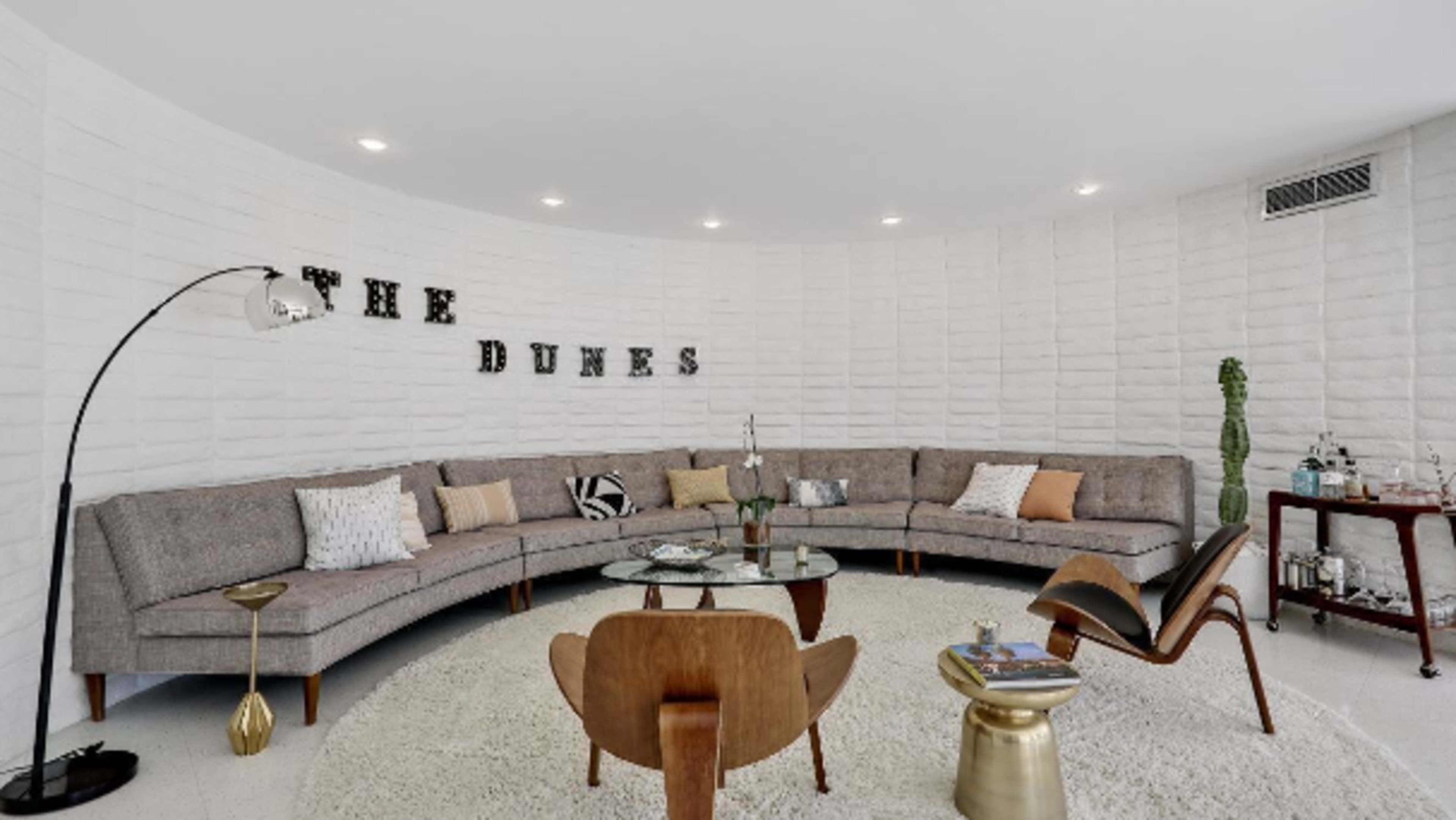 A curved living room features a large sectional sofa, a round glass table, and decorative elements against a white brick wall with "THE DUNES" displayed prominently.