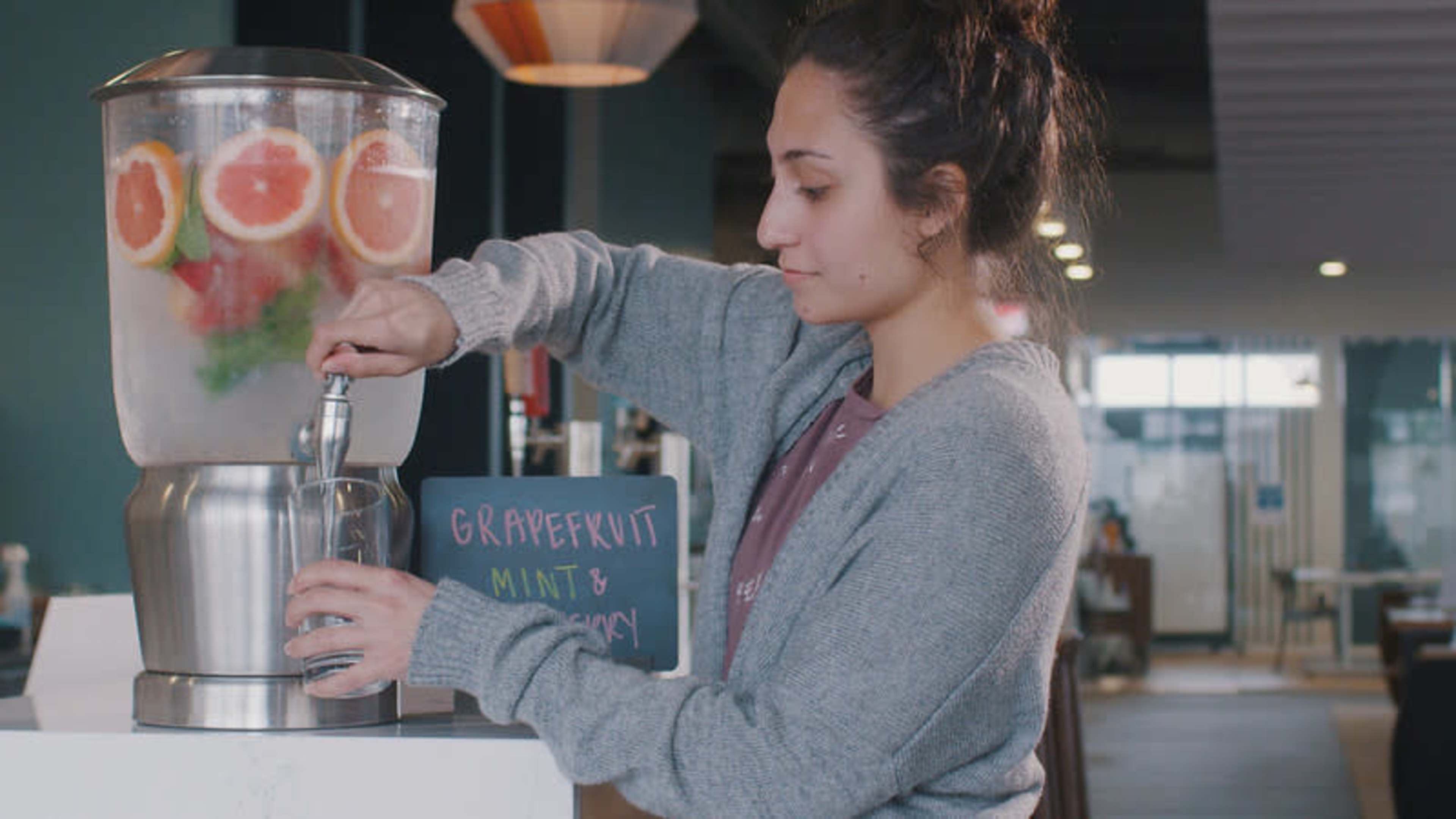 A woman is filling a glass with infused water from a beverage dispenser containing grapefruit and mint.