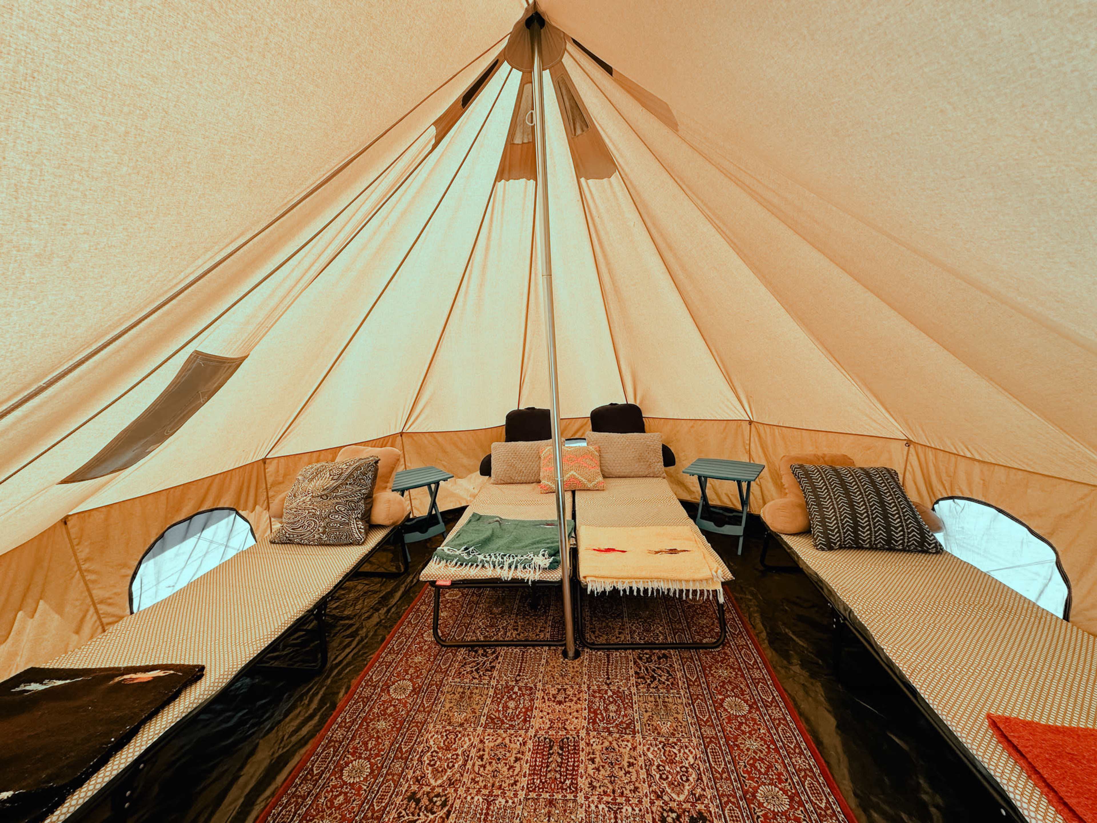 A spacious tent interior features two neatly arranged cots with decorative pillows and a central rug.