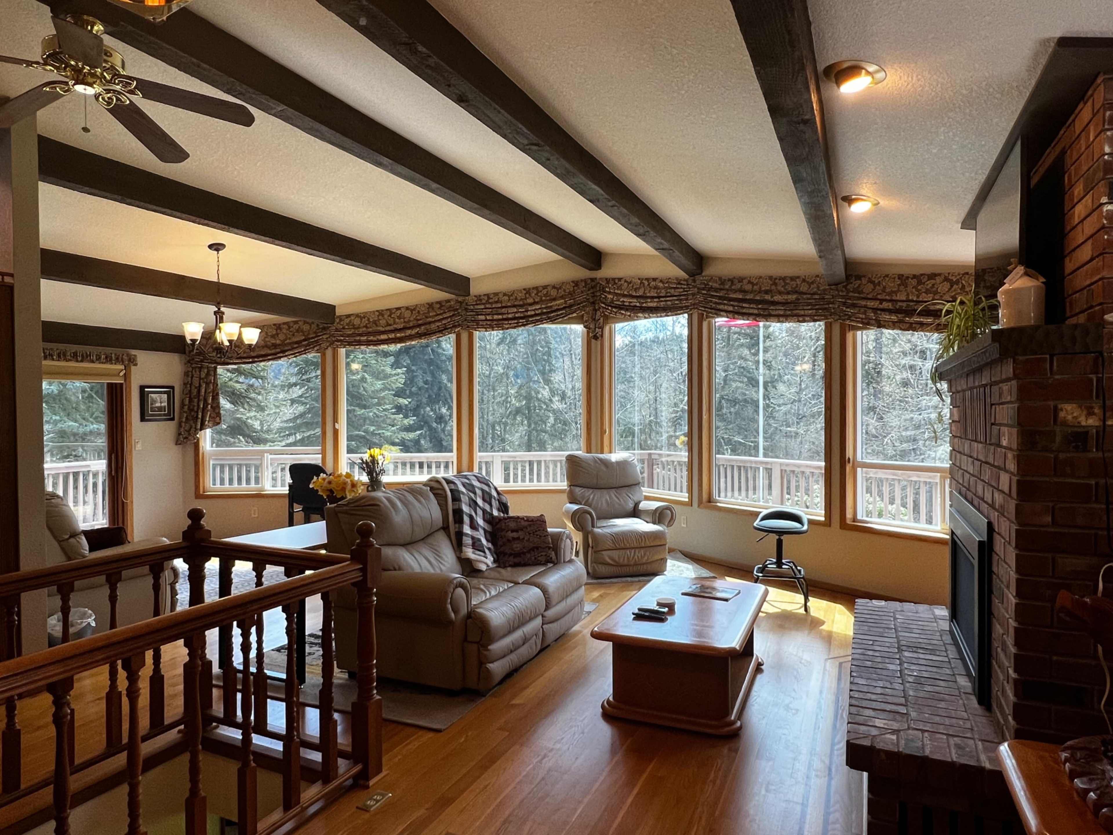 The image shows a living room with large windows overlooking a wooded area, featuring beige leather furniture and a brick fireplace.