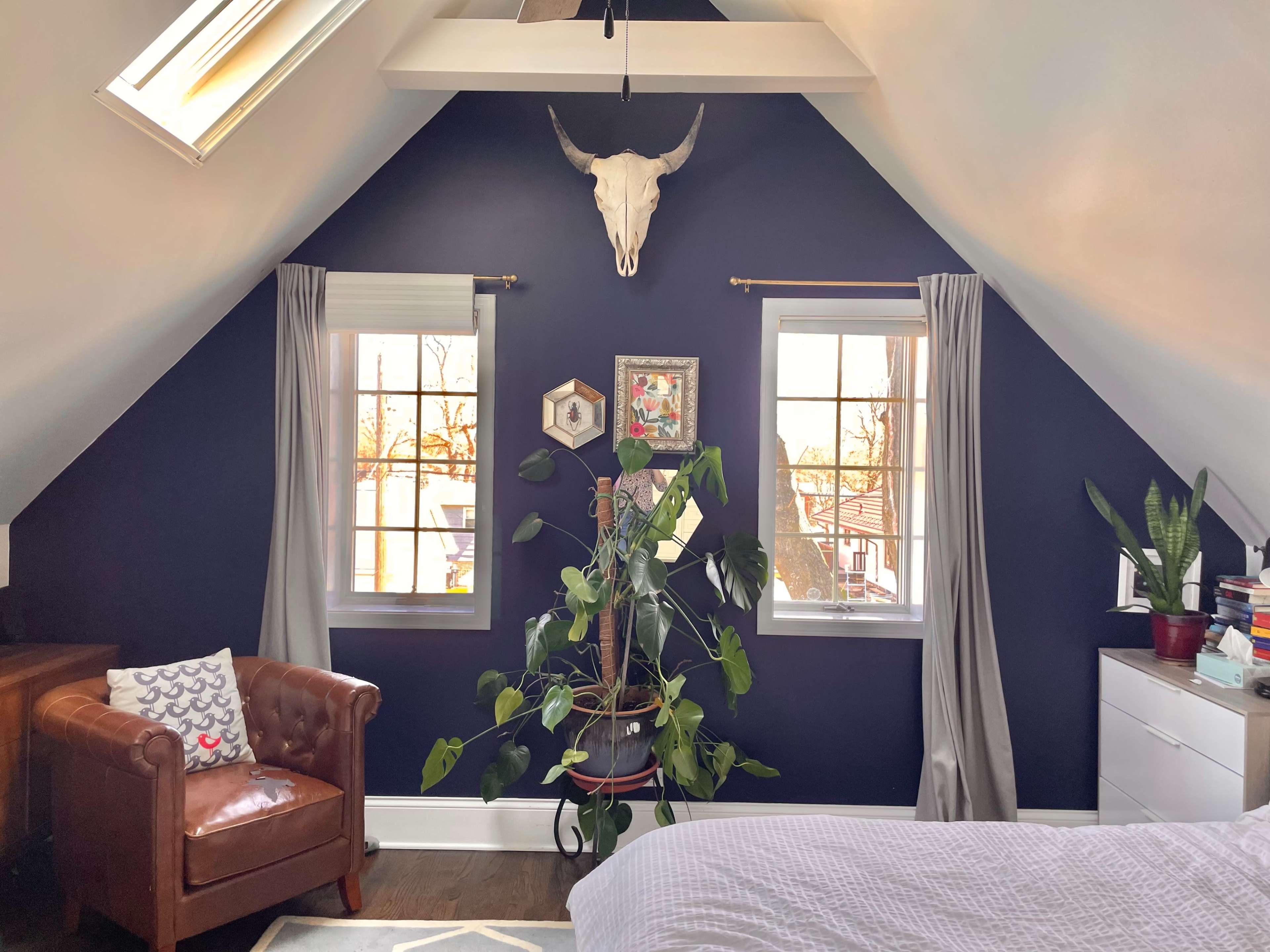 An attic room with sloped walls, featuring a brown leather chair, large windows, a plant, and a cow skull mounted on the wall.