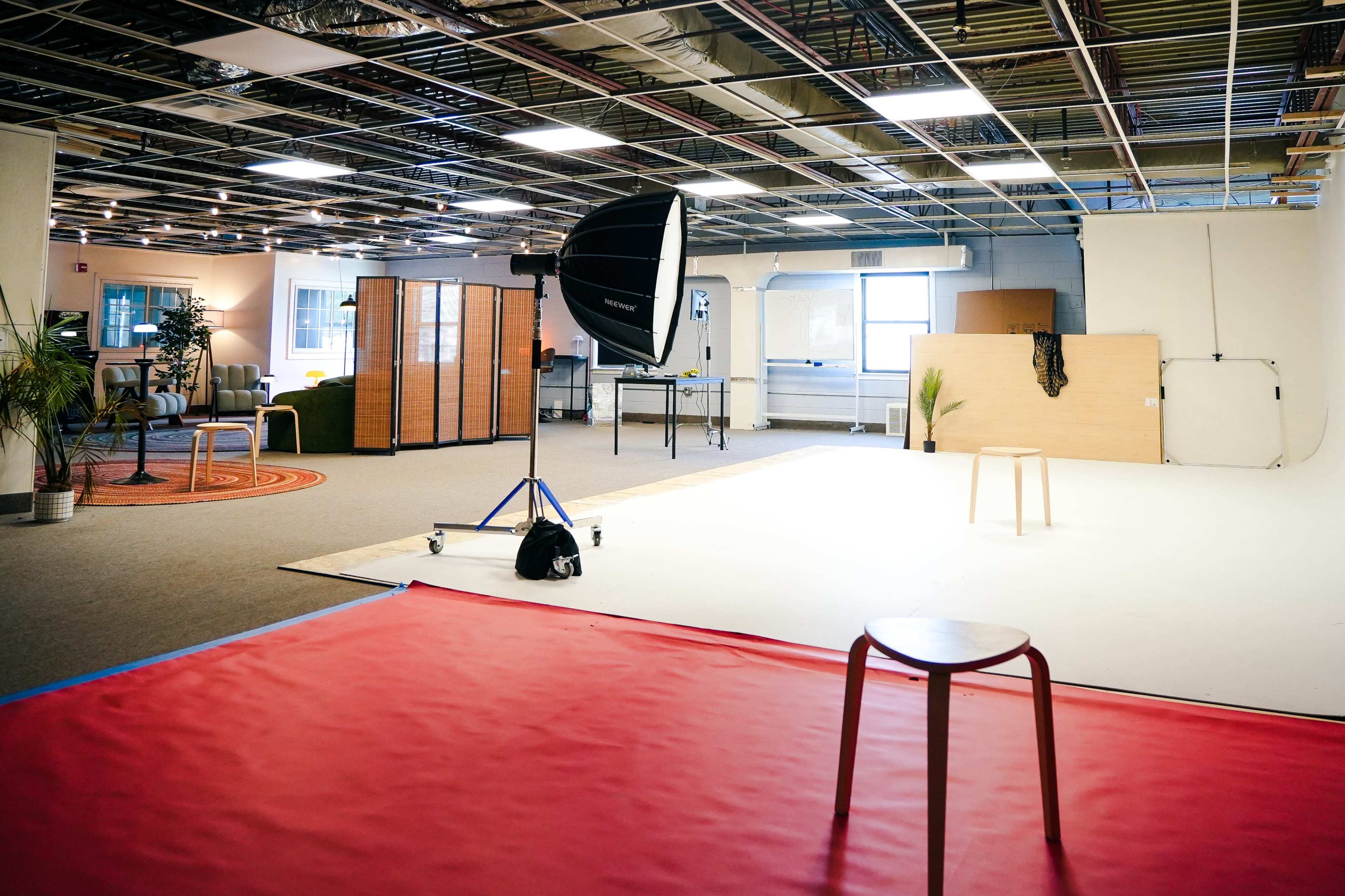 The image shows a spacious studio with a red mat, a stool in the center, lighting equipment, and partitions creating distinct areas.