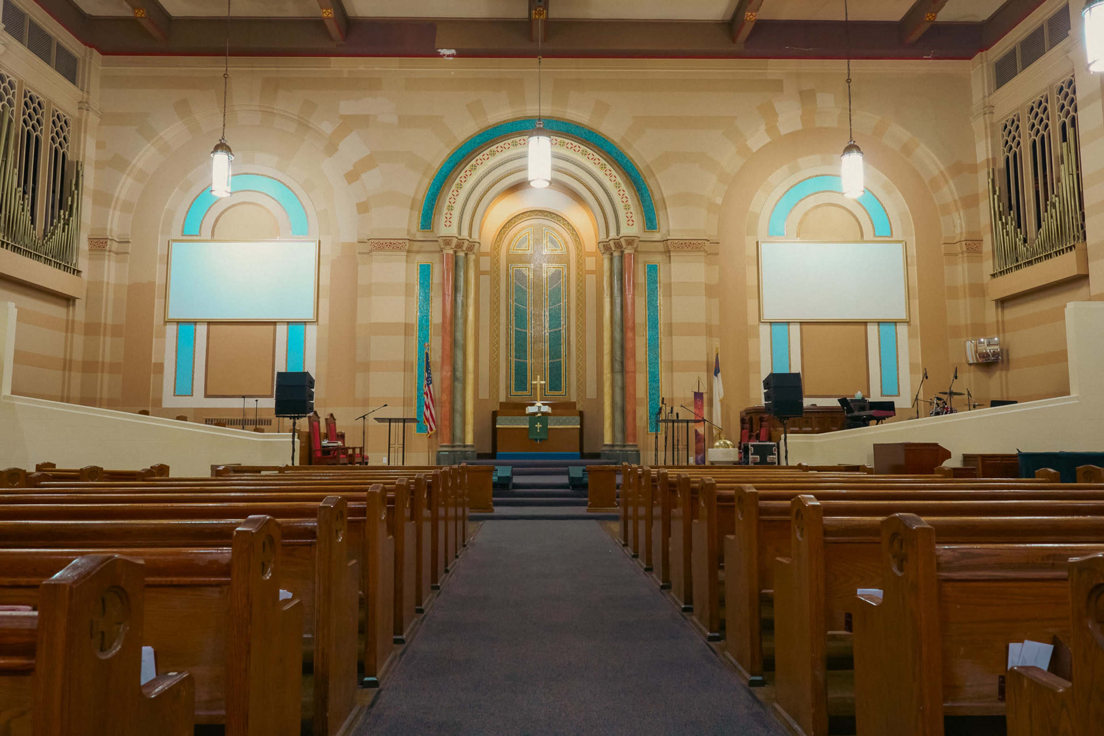 The image shows the interior of a church with wooden pews facing a raised altar area, illuminated by hanging lights and large windows.