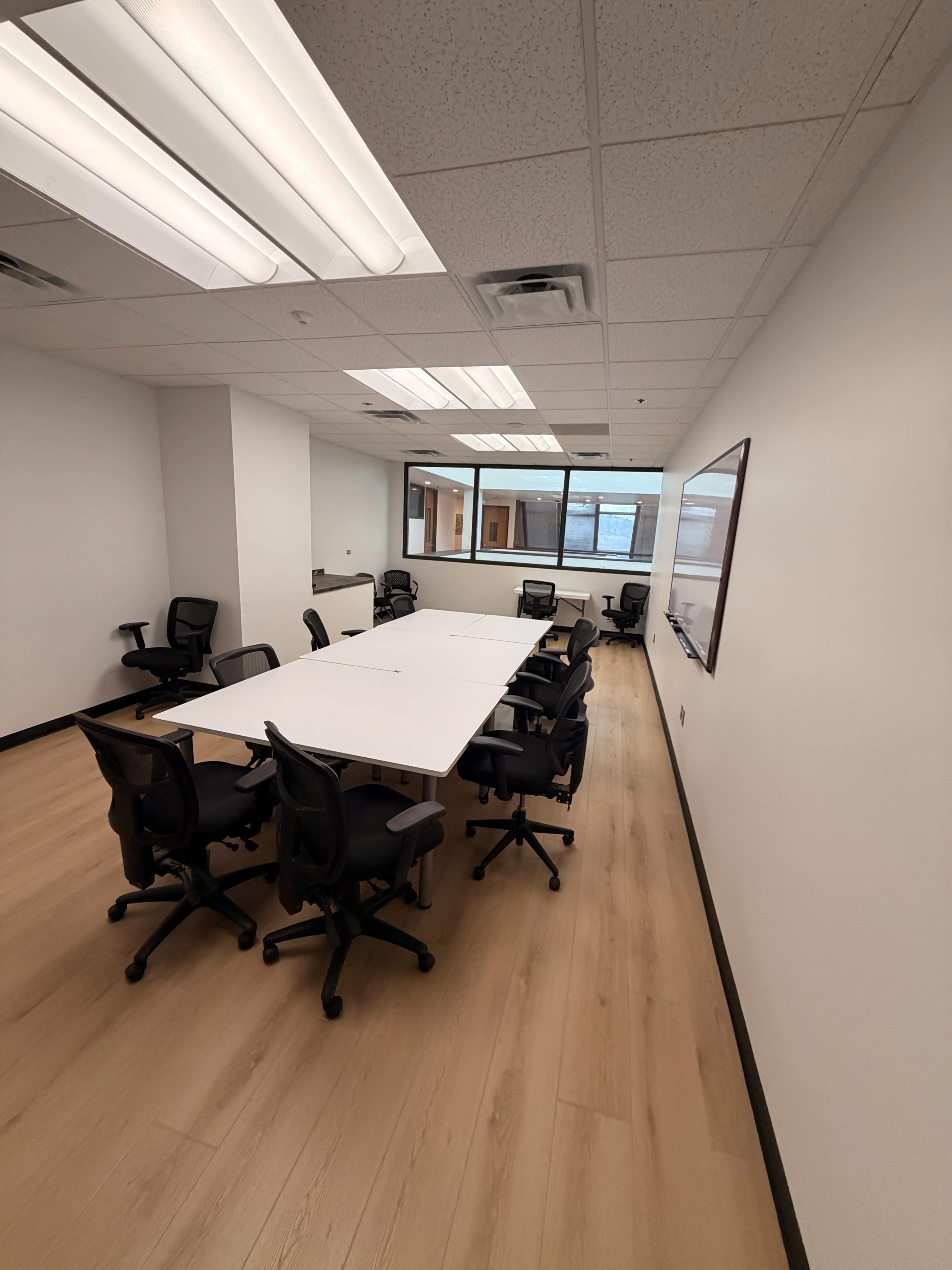The image shows a modern conference room with a long table surrounded by black office chairs and large windows providing natural light.