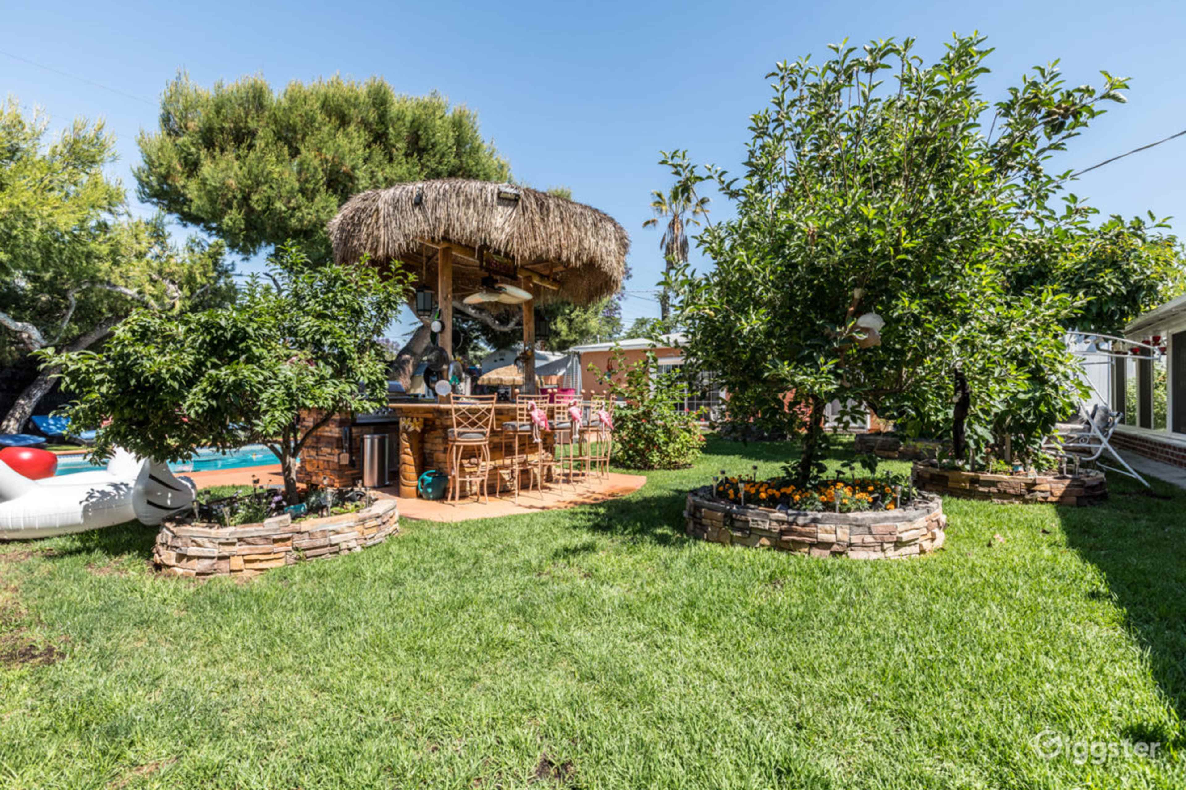 A tropical-themed outdoor bar with a thatched roof surrounded by landscaped greenery and garden features.