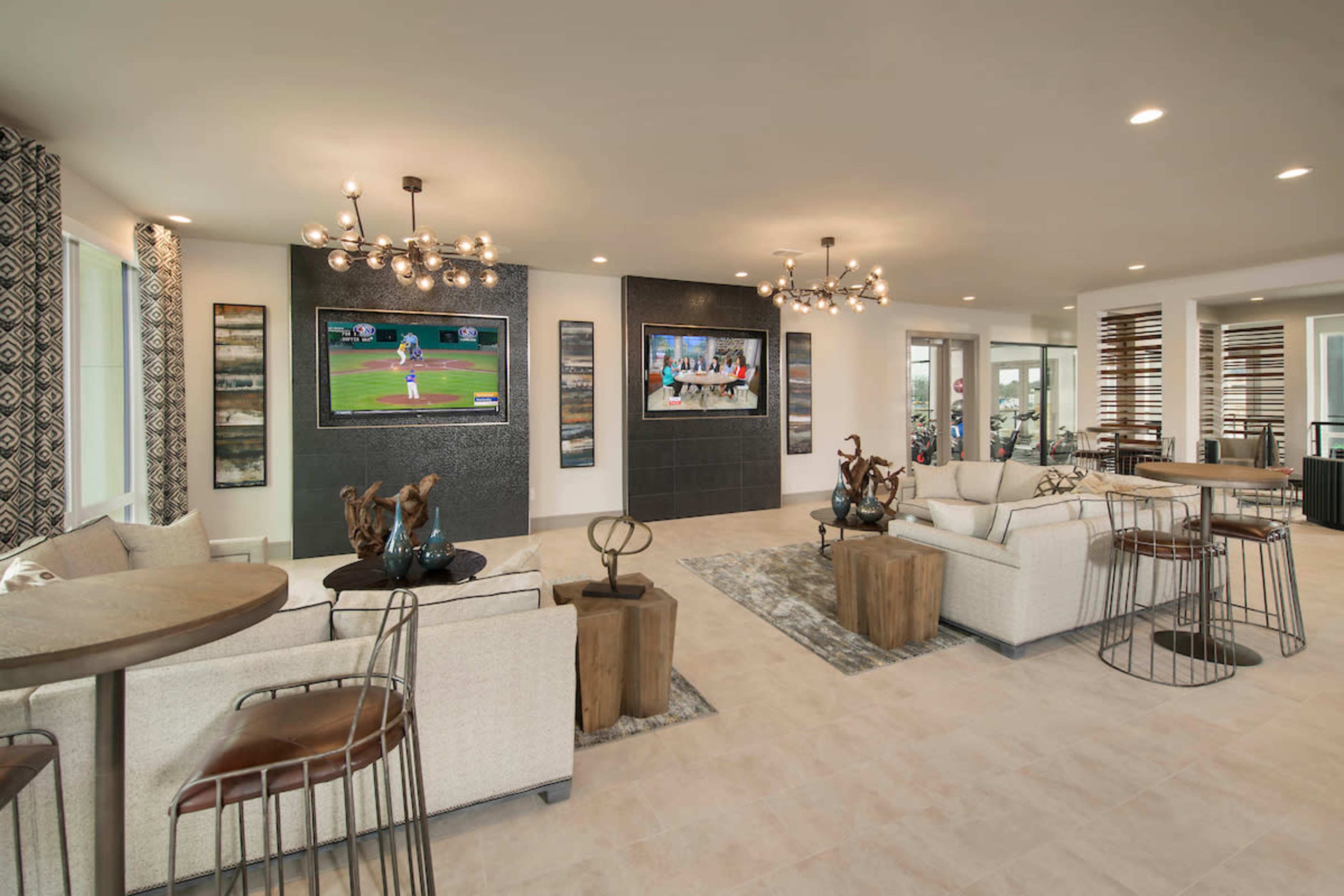 A modern living space with two televisions mounted on the wall, seating areas with light-colored sofas, and several decorative tables featuring sculptures.
