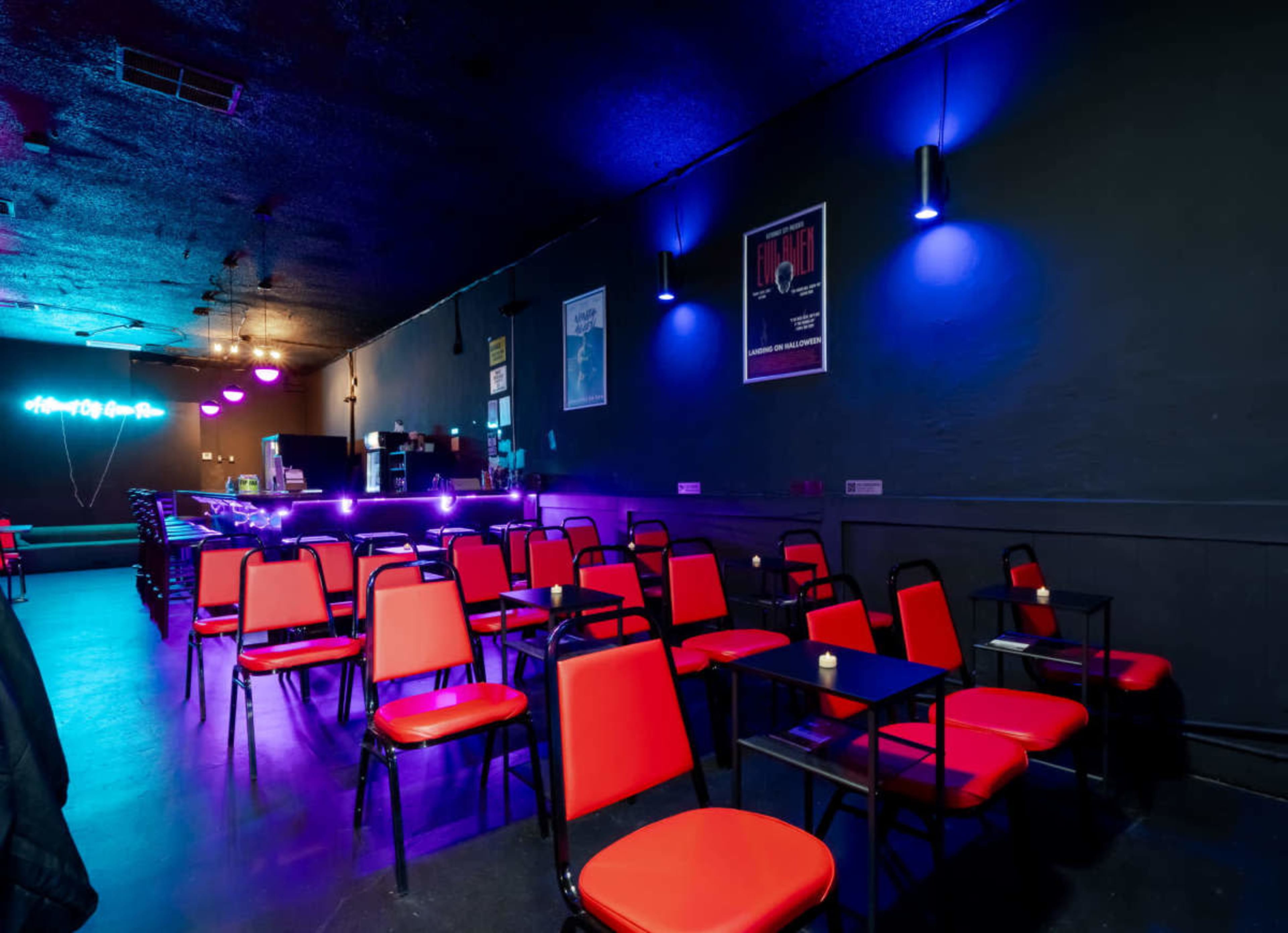 A dimly lit lounge features red chairs and tables arranged neatly, with a bar in the background illuminated by purple and blue lights.