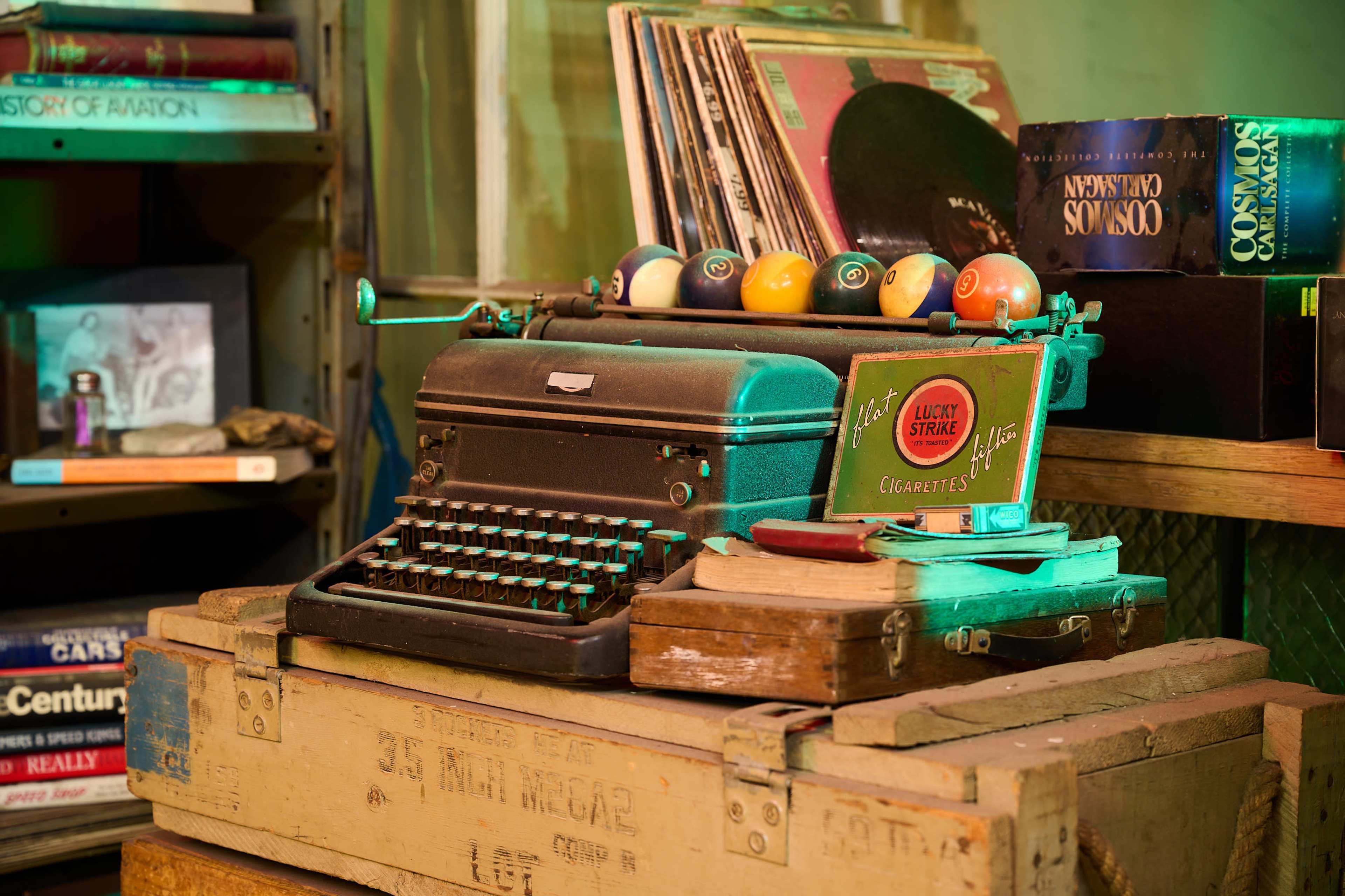A vintage typewriter sits atop a stack of wooden crates alongside a set of colorful billiard balls and an old cigarette tin in a dimly lit room.