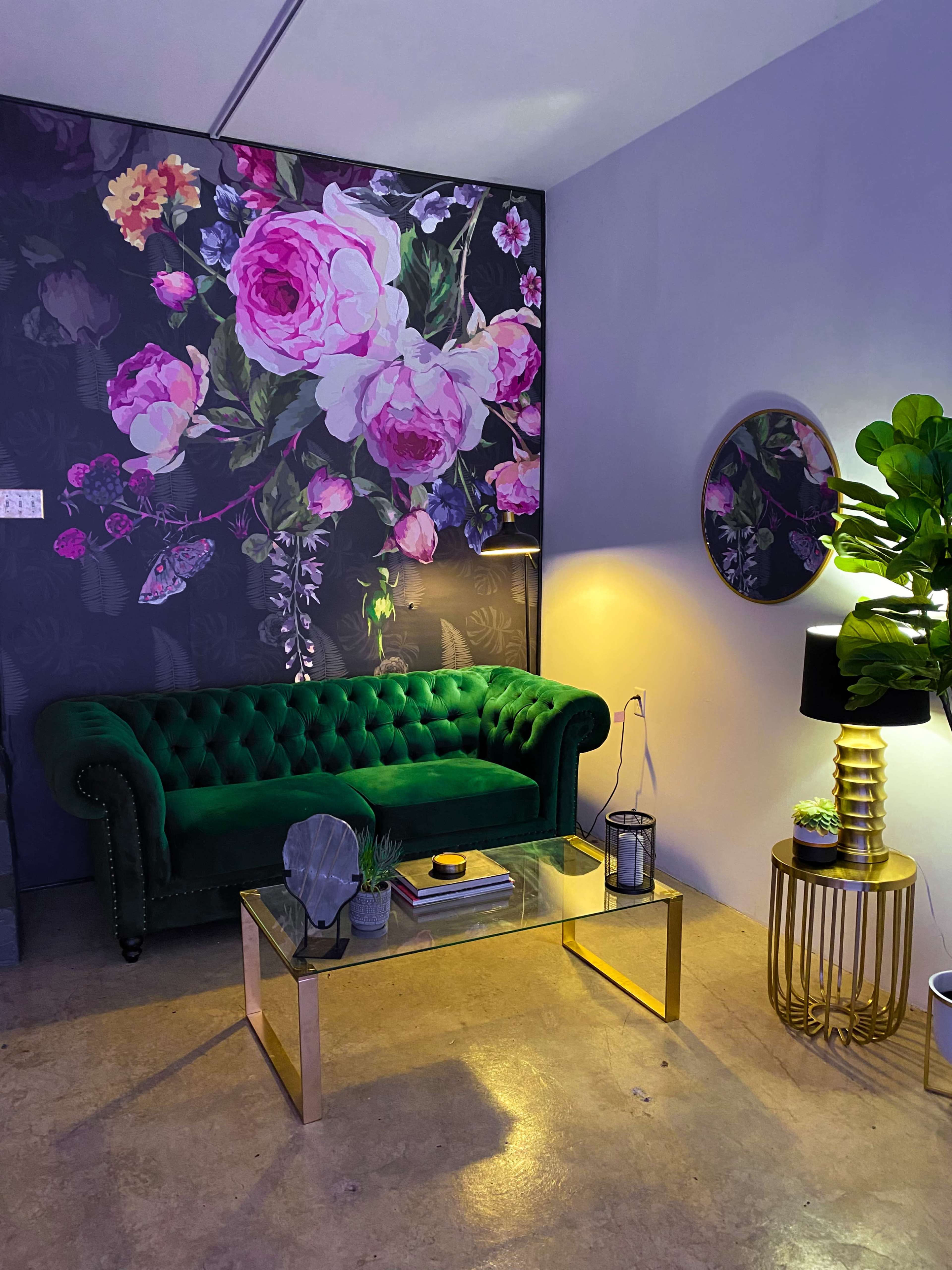 The scene features a green velvet tufted sofa against a floral wall mural, accompanied by a glass coffee table and decorative elements including a plant, lamp, and wall mirror.