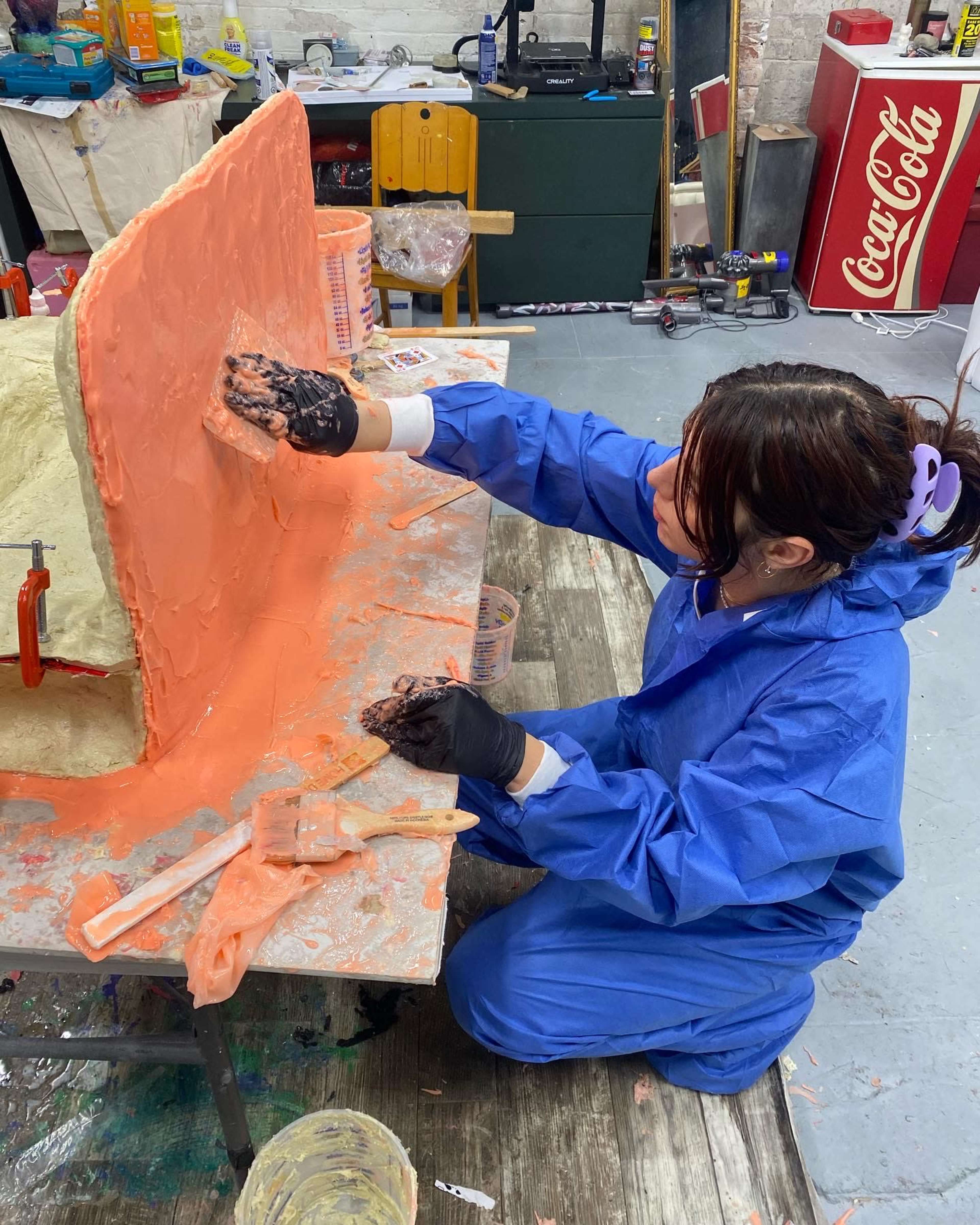 A person in a blue jumpsuit is applying orange material to a mold on a workbench in a workshop.