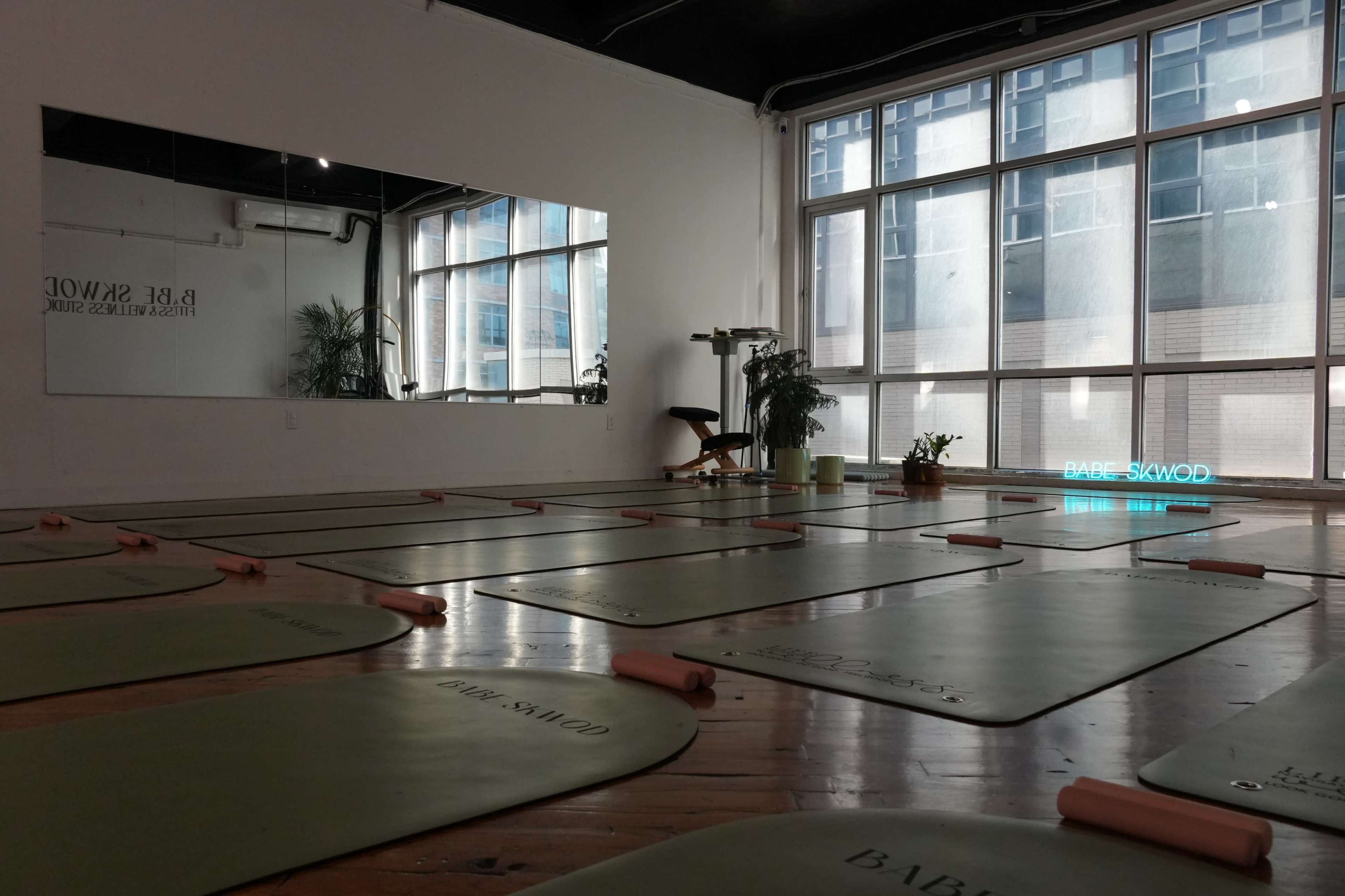 The image shows a yoga studio with several exercise mats arranged on a wooden floor, a large mirror along one wall, and large windows allowing natural light to enter.