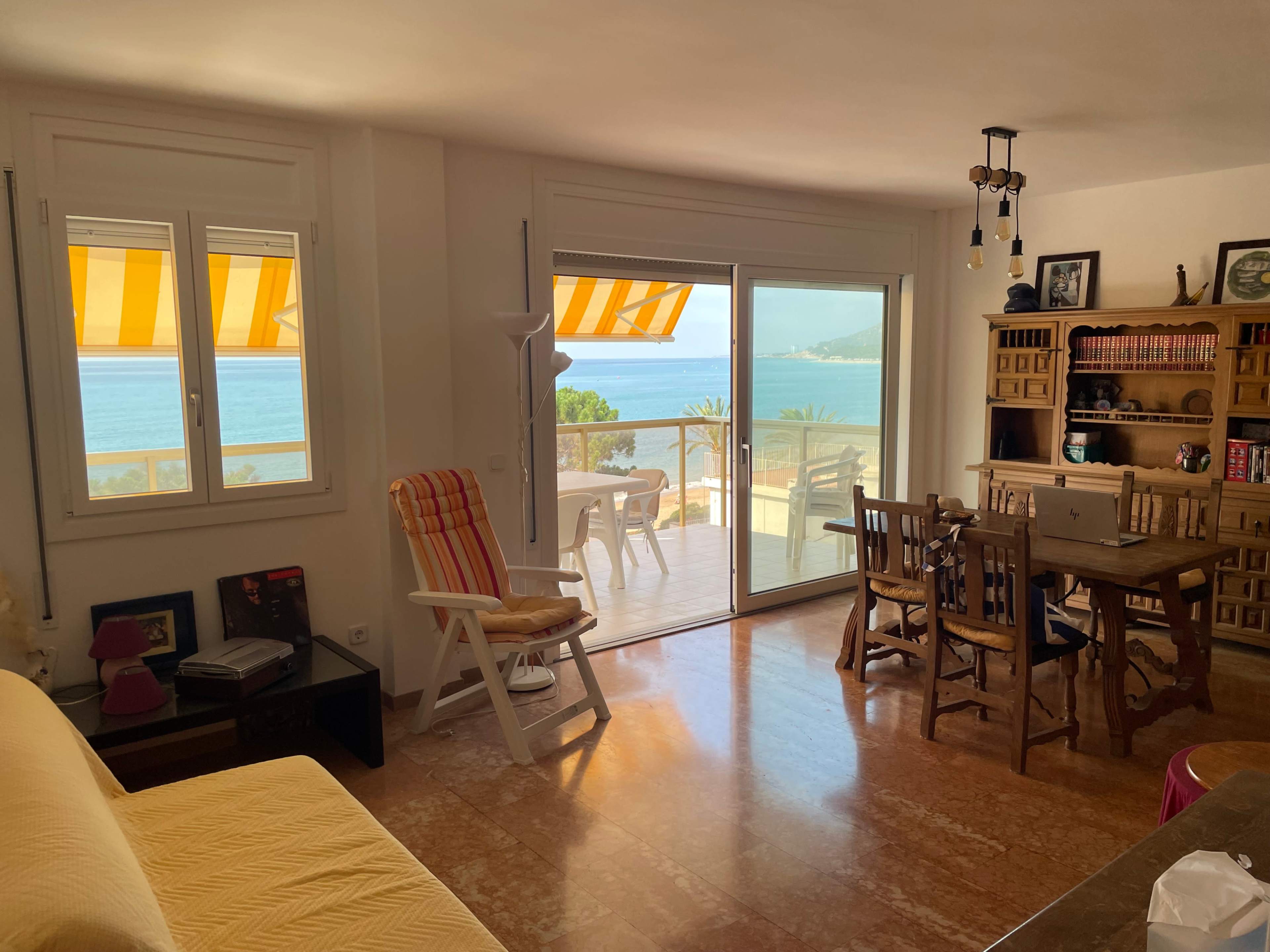 The image shows a bright living space with a view of the ocean through large glass doors, featuring a dining area and comfortable seating.