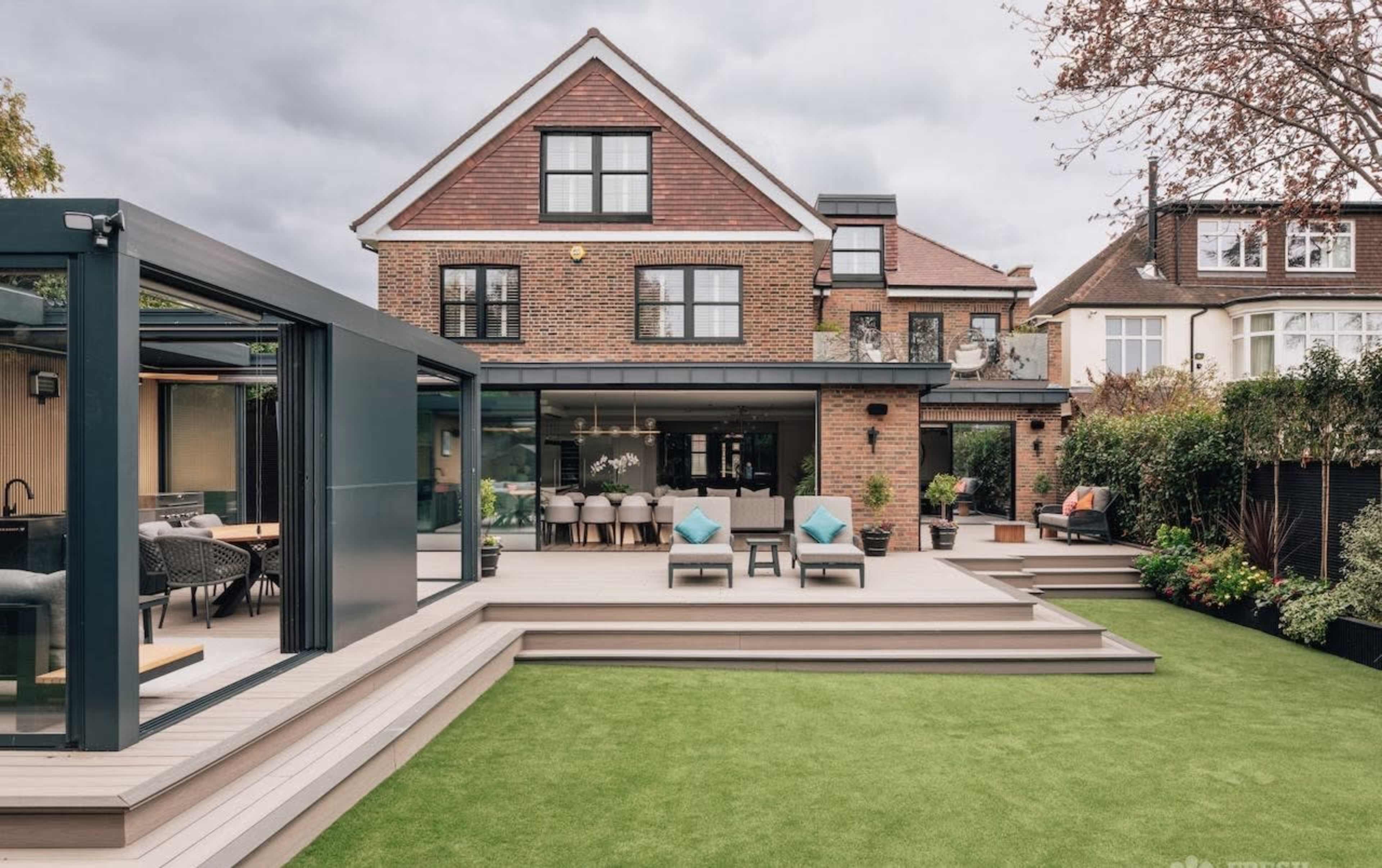 The image shows a modern backyard with a well-maintained lawn, a multi-level deck, and a large house featuring a glass extension and outdoor seating areas.