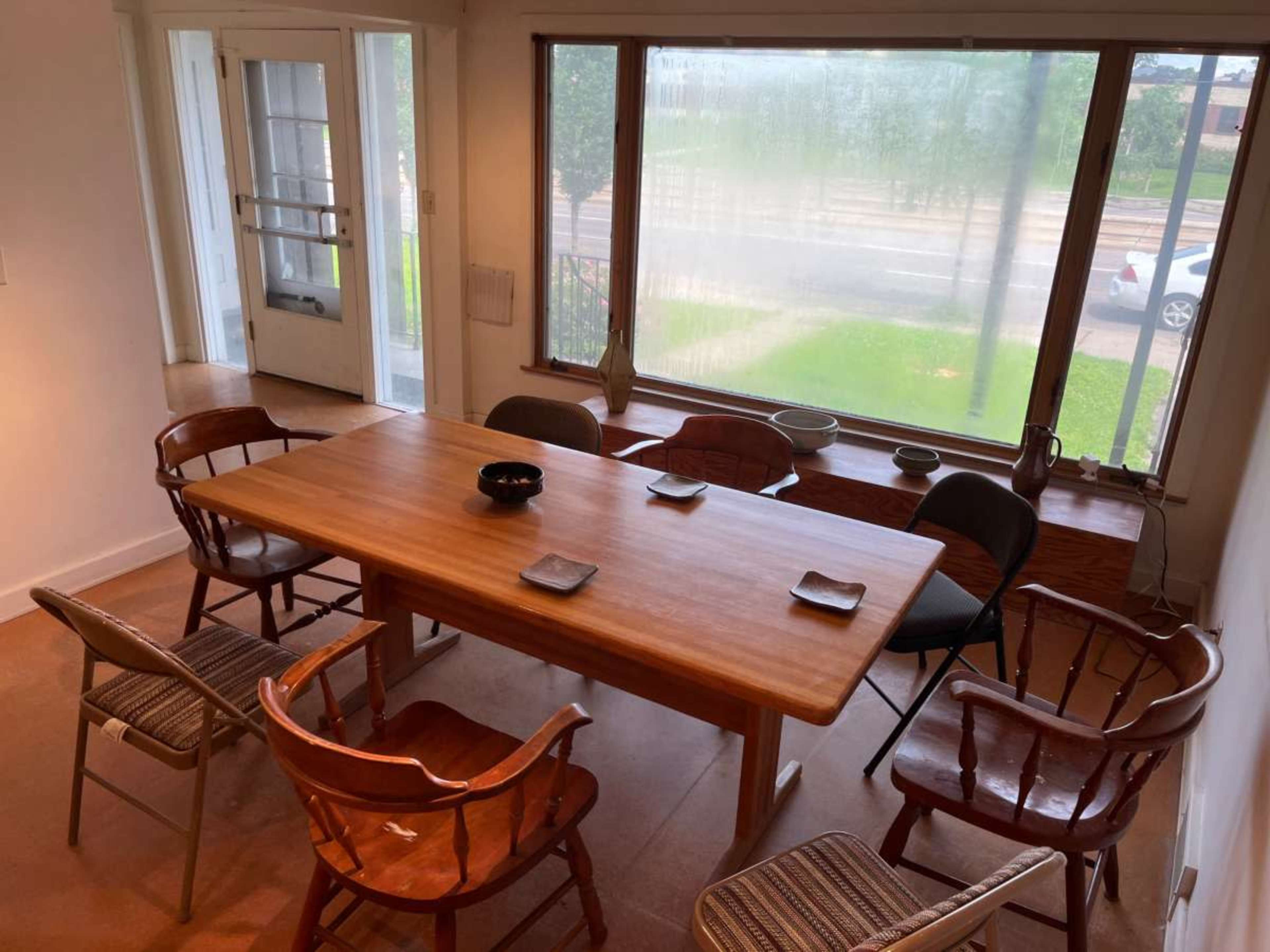 A wooden dining table surrounded by eight chairs is positioned in a room with large windows, revealing a view of the outside.