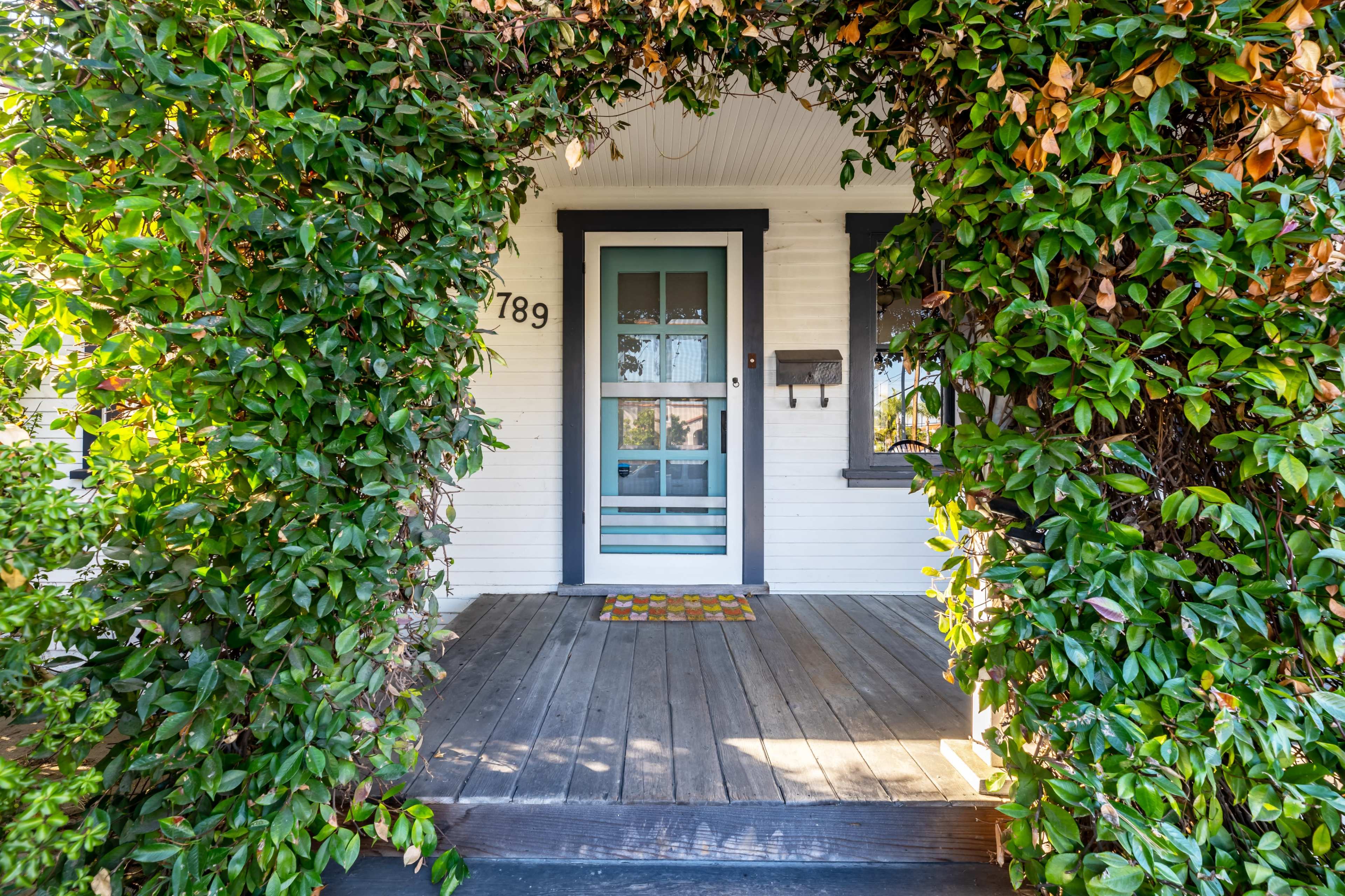 The image shows a porch entrance framed by green foliage, leading to a blue door with a welcome mat on a wooden floor.