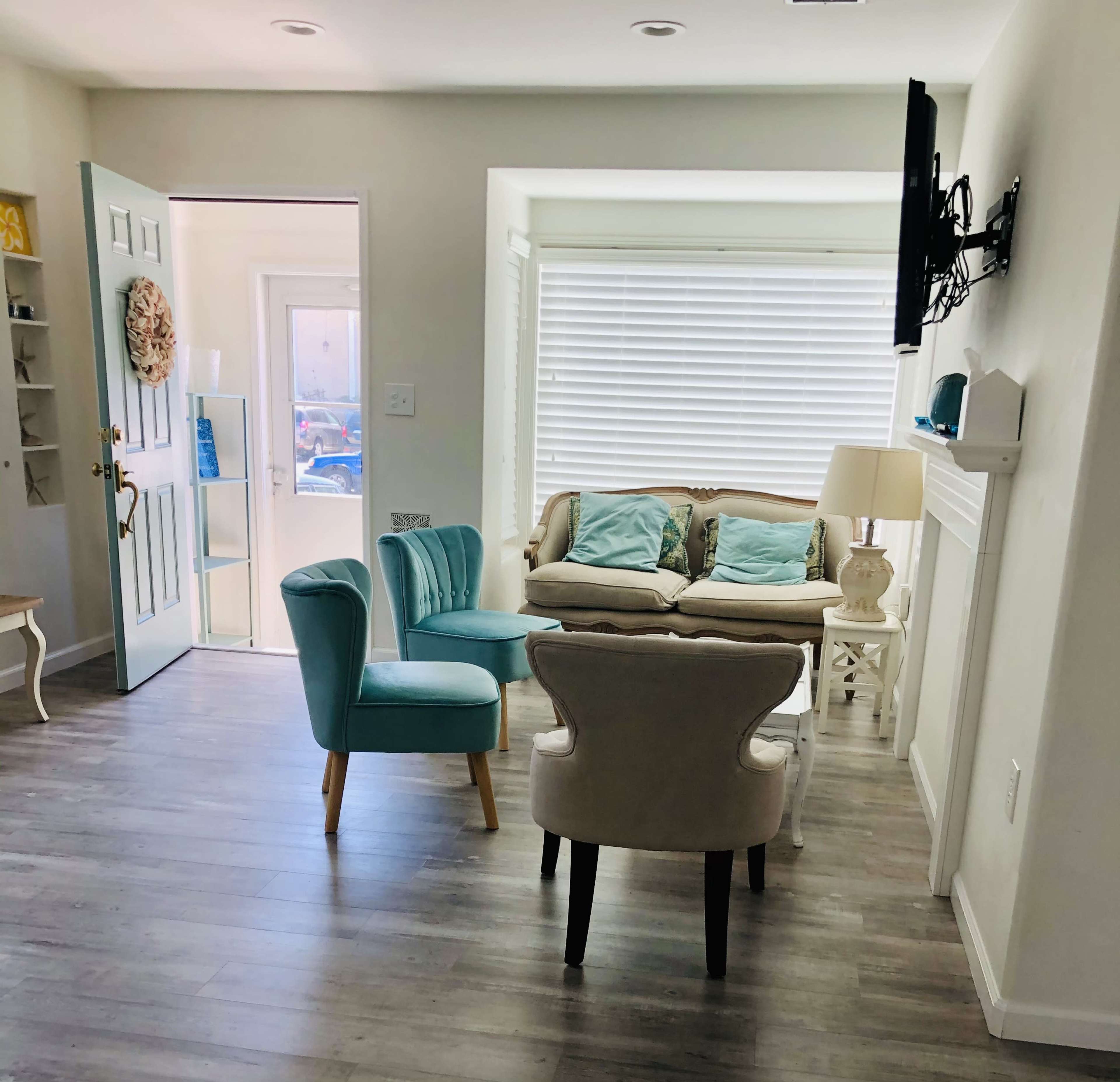 A bright living room features a blue chair, a cream-colored chair, and a small sofa, with a door leading outside and a television mounted on the wall.