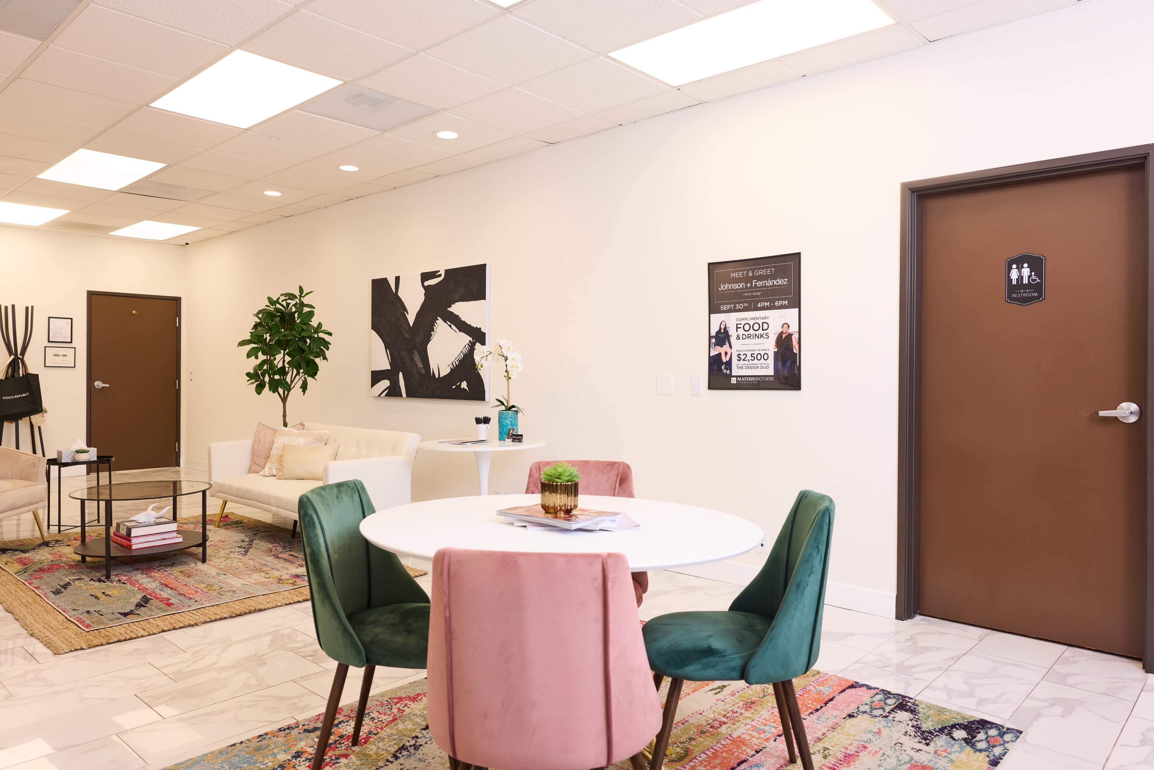 A modern waiting area with a round dining table surrounded by four chairs, a seating area with a sofa, and a restroom door on the right side.