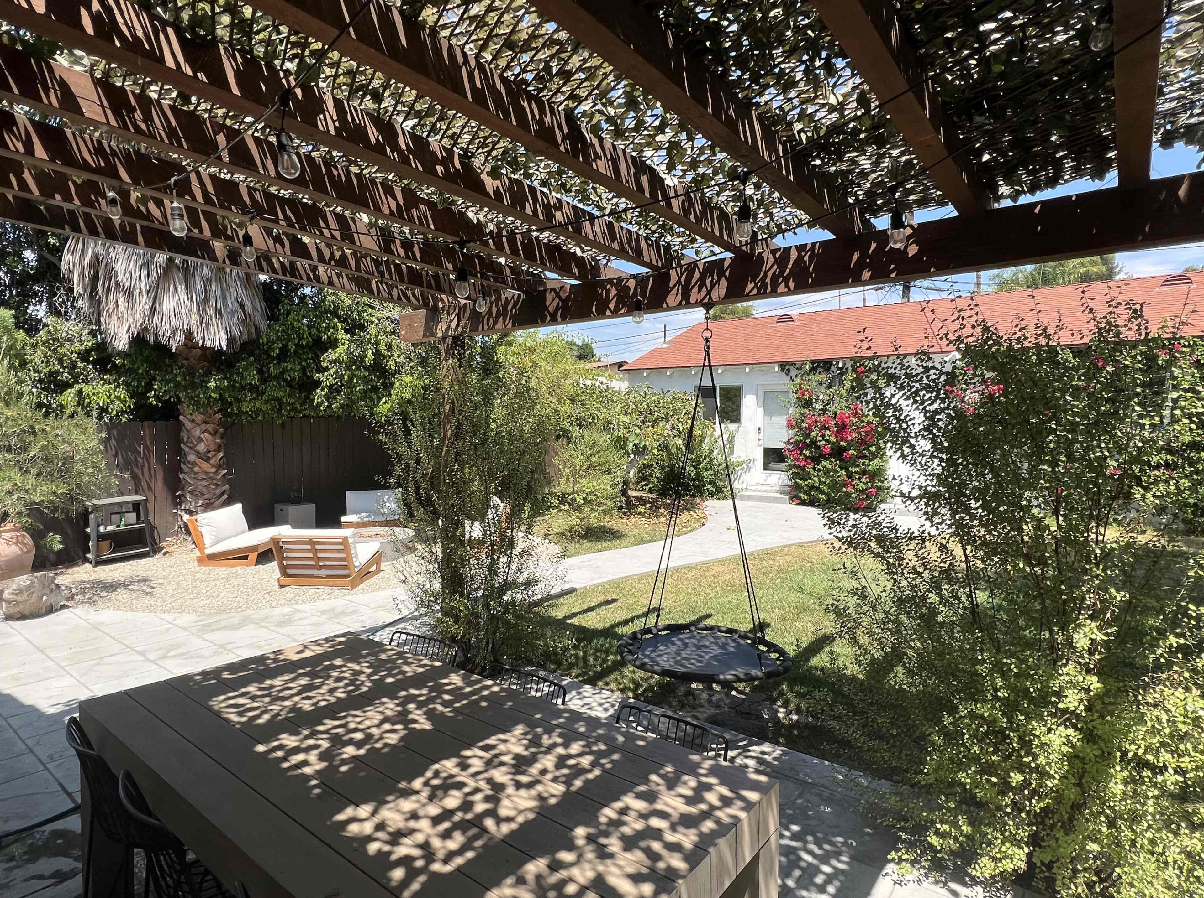 A shaded patio with a wooden table overlooks a landscaped yard featuring a swing and lounge chairs.