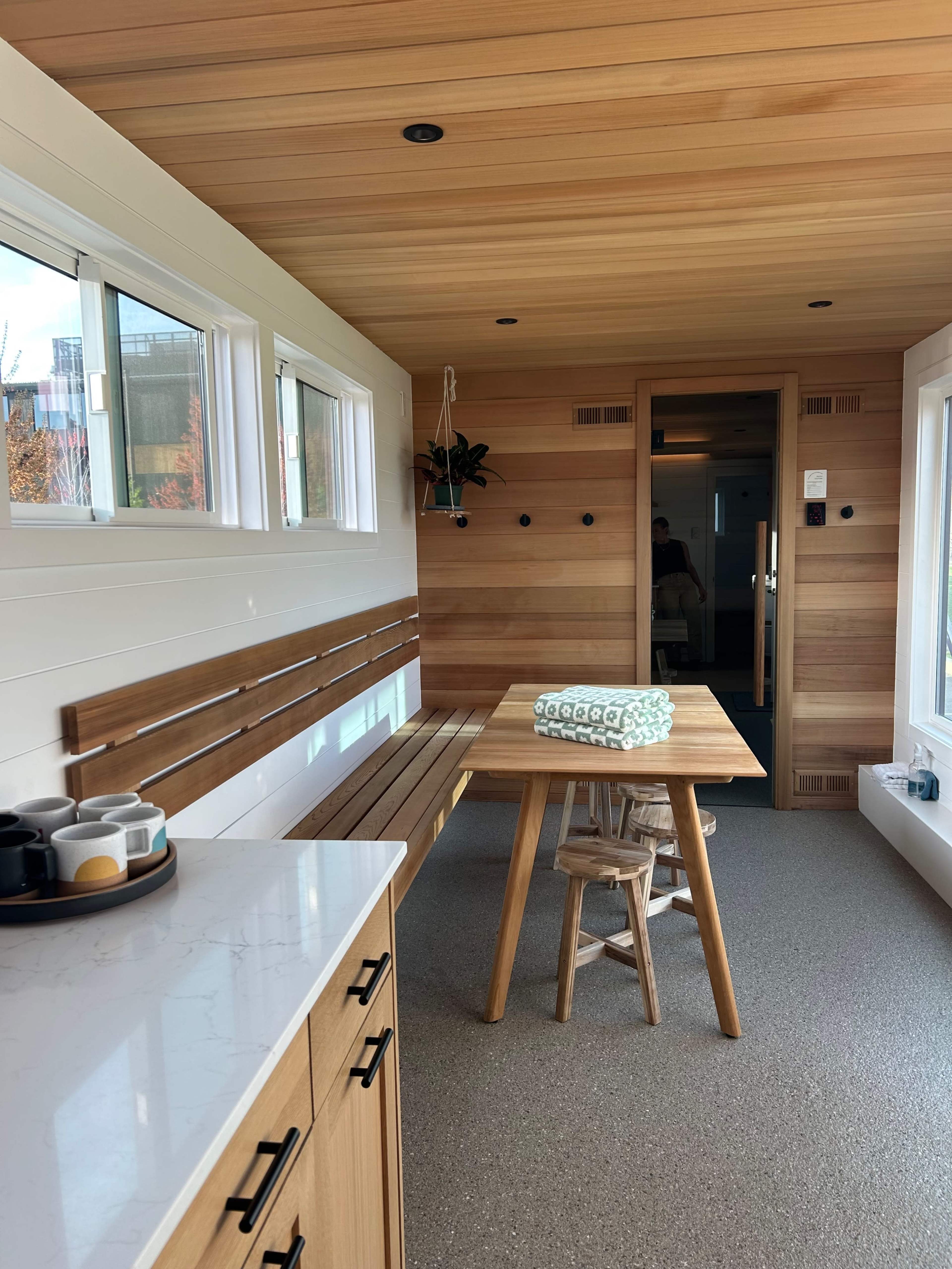 The image shows a modern, wooden interior space featuring a dining table with a small chair, a bench along one wall, and a kitchen area with a countertop.