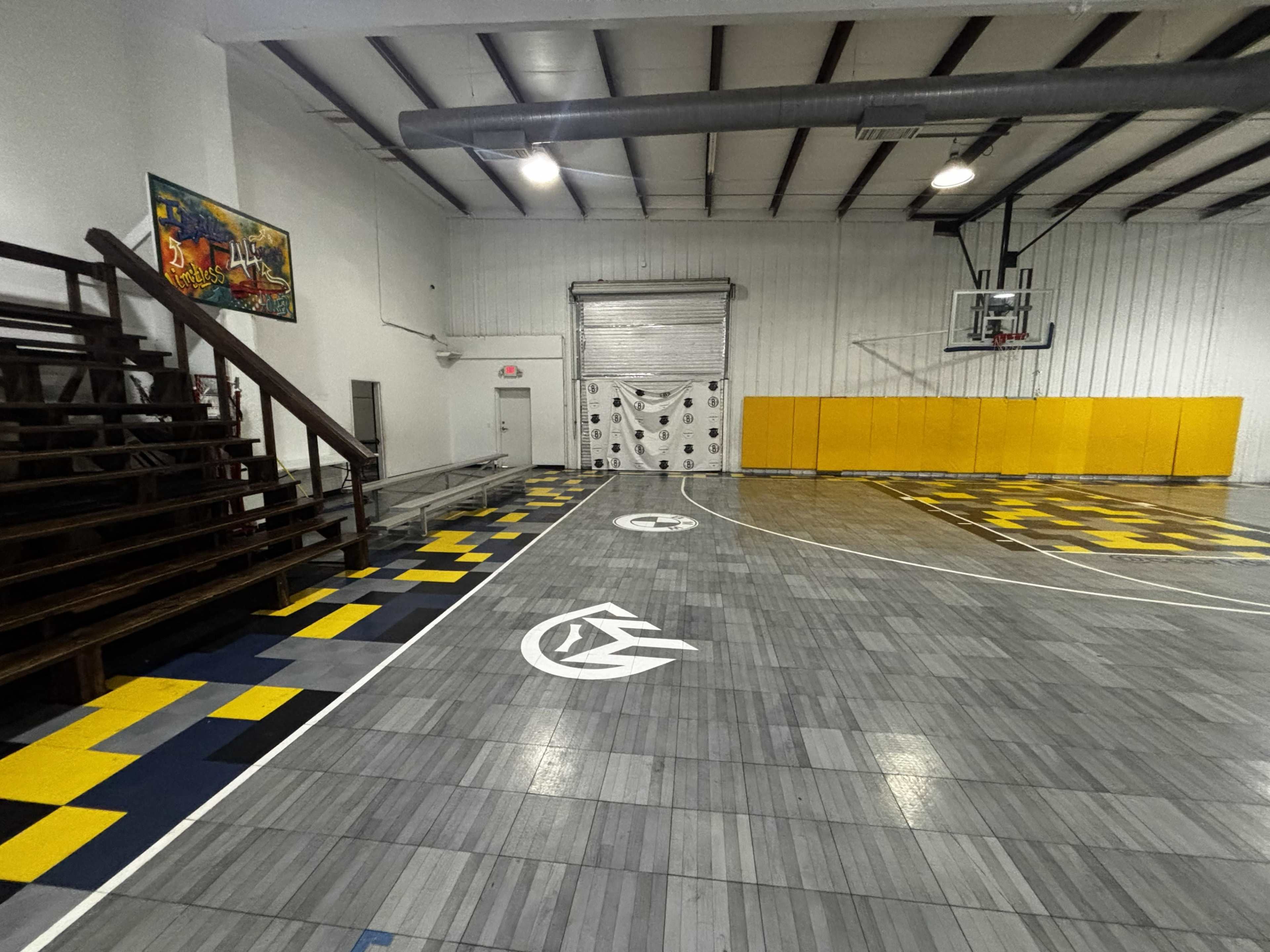 The image shows a basketball court with a unique patterned floor, featuring yellow and gray tiles, bleacher seating on the left, and a basketball hoop in the background.