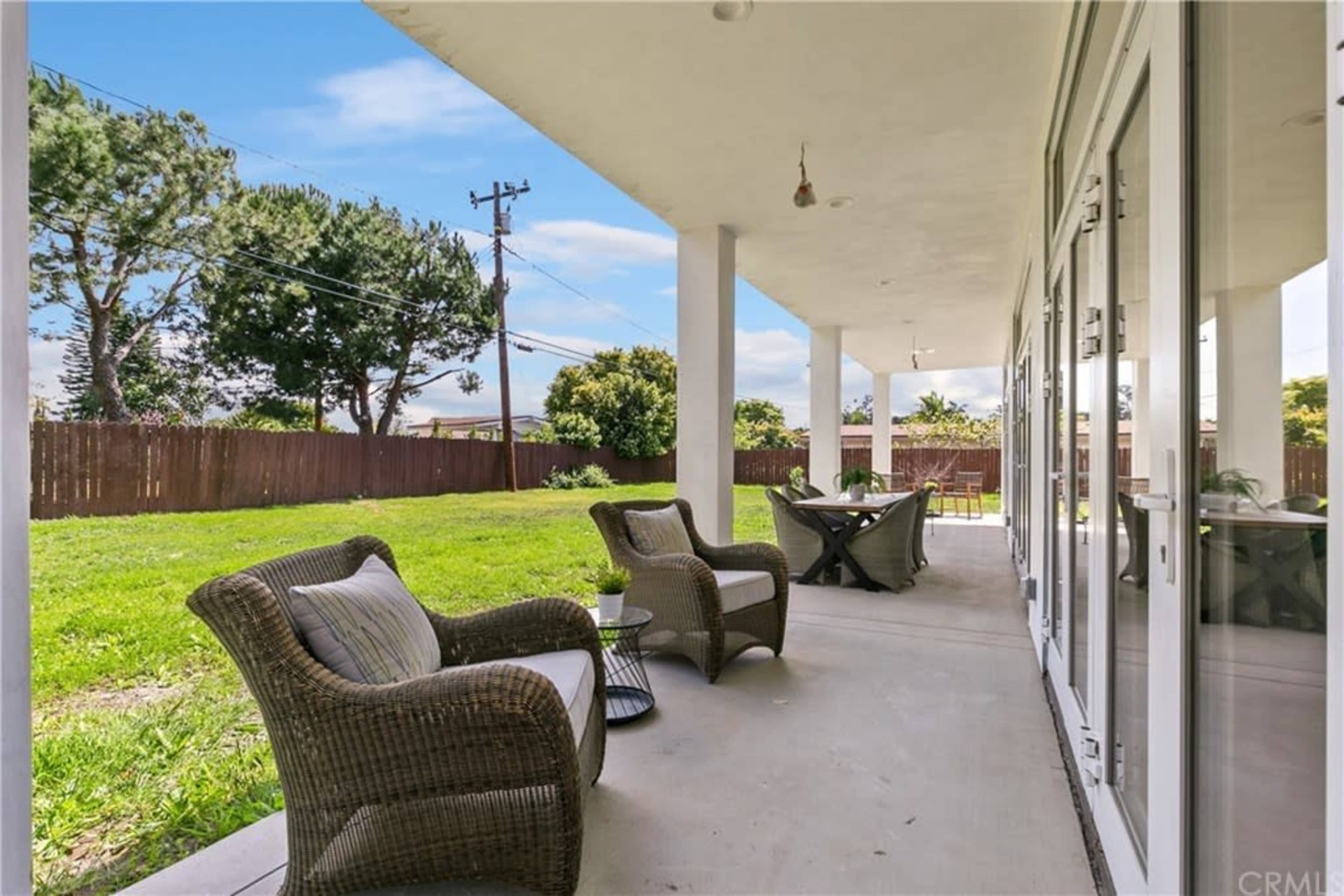 A covered patio features two chairs and a table, overlooking a grassy yard with trees in the background.