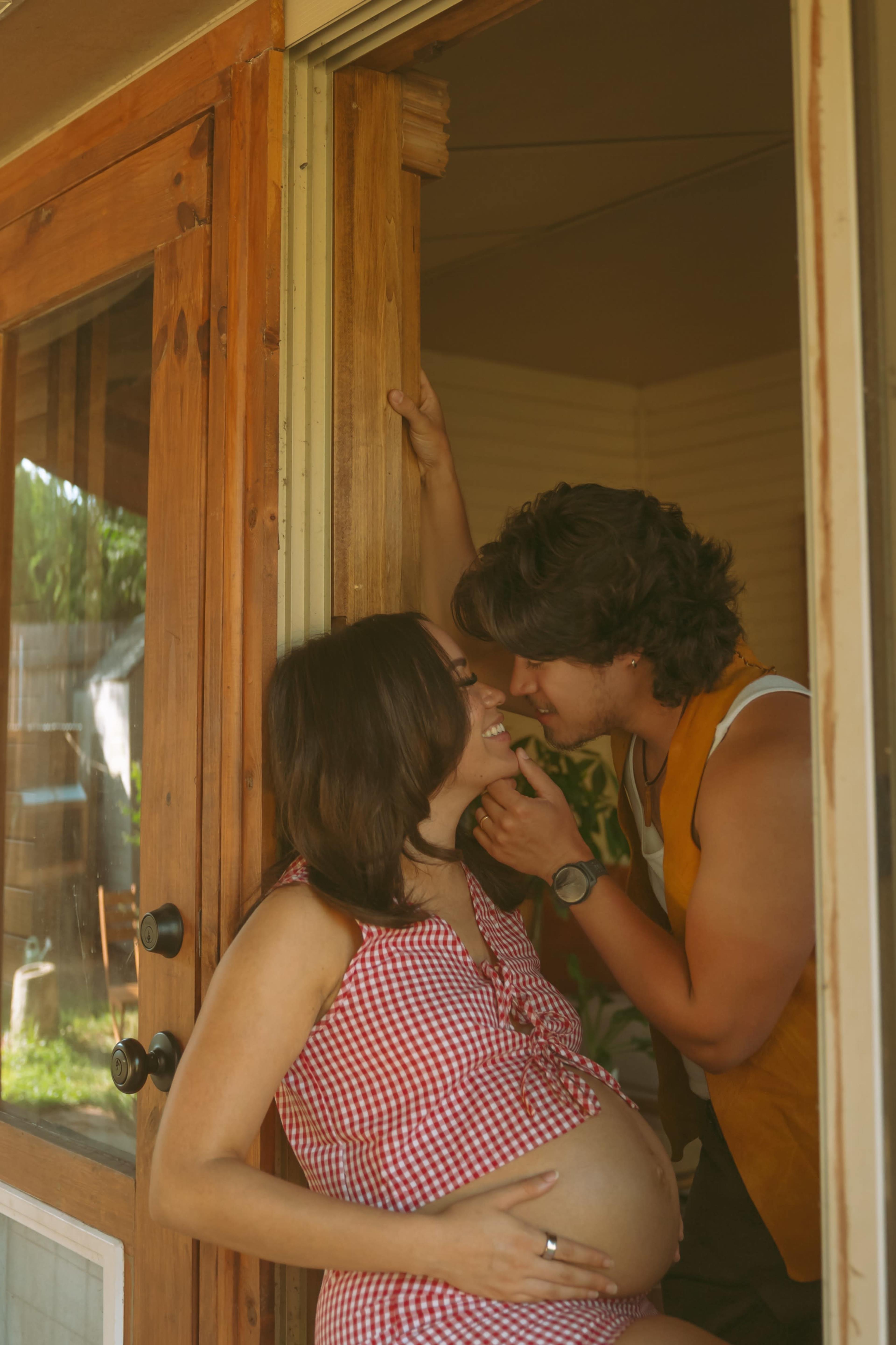 A couple shares a tender moment at a doorway, with the pregnant woman in a checkered dress and the man leaning in close.
