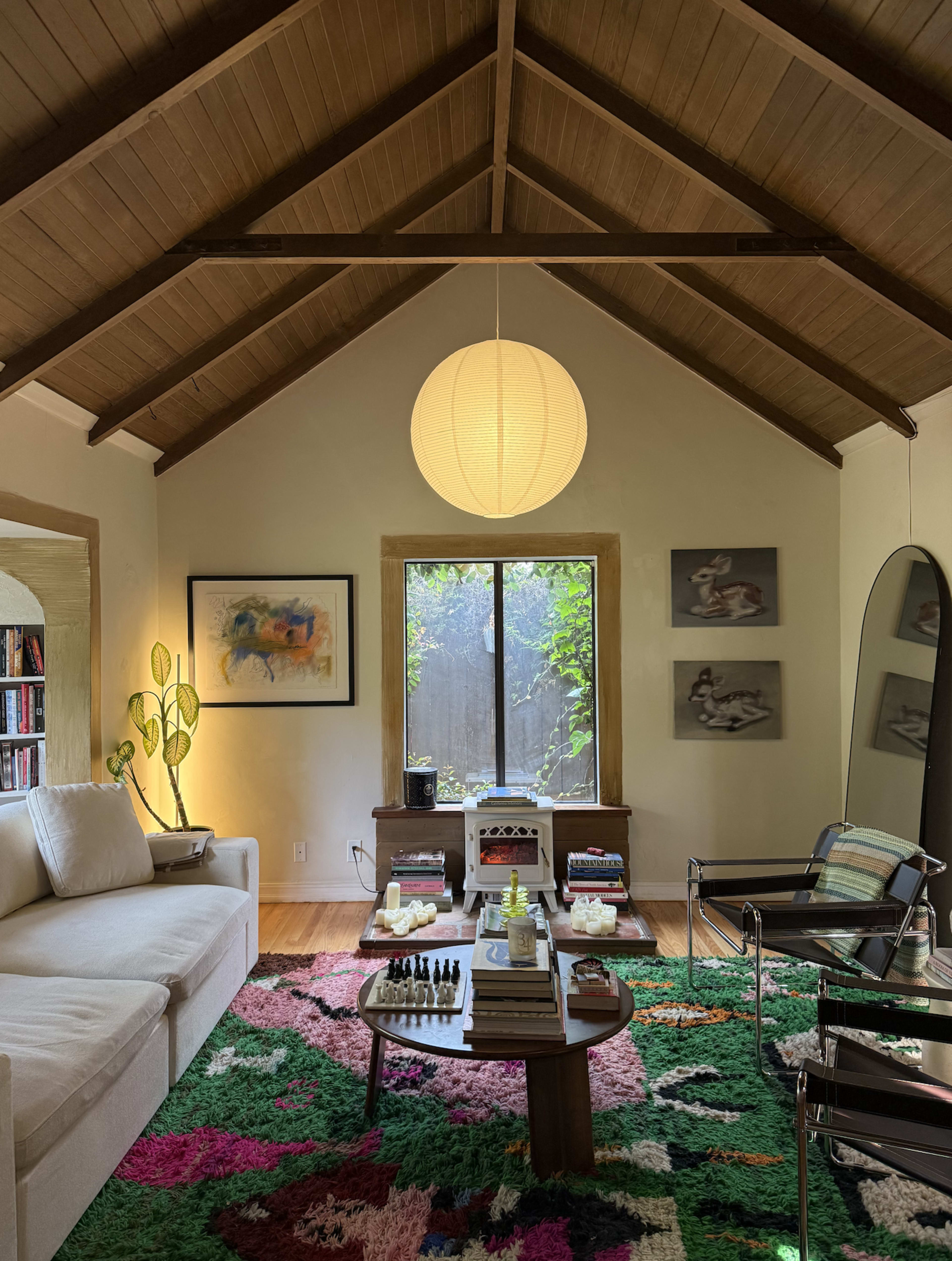 The image shows a cozy living room with a high, wooden ceiling, featuring a large window, a circular pendant light, and colorful rugs with a modern seating arrangement.