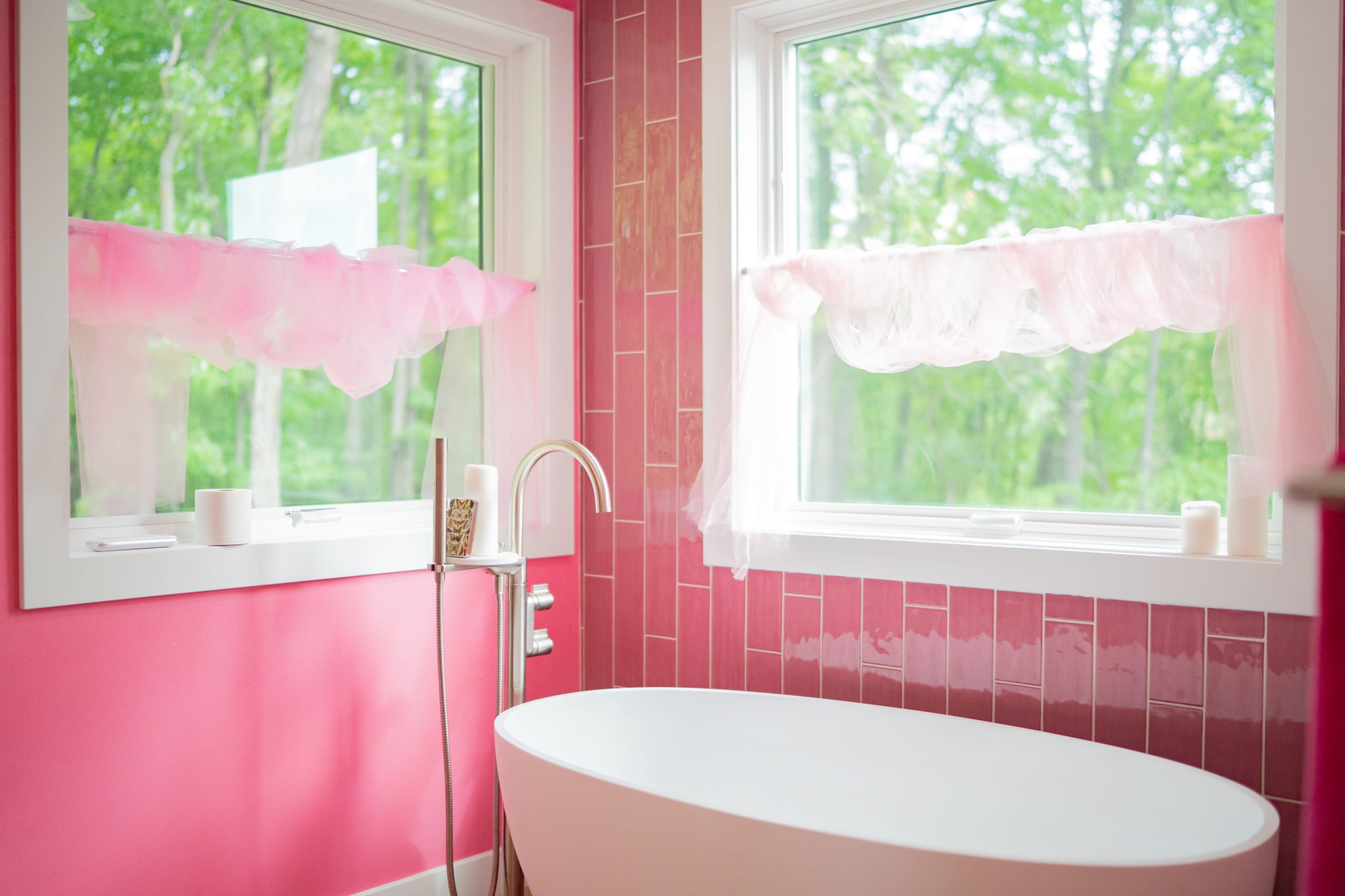 The image shows a bright pink bathroom with a freestanding white bathtub and large windows draped with sheer fabric, overlooking a green wooded area.