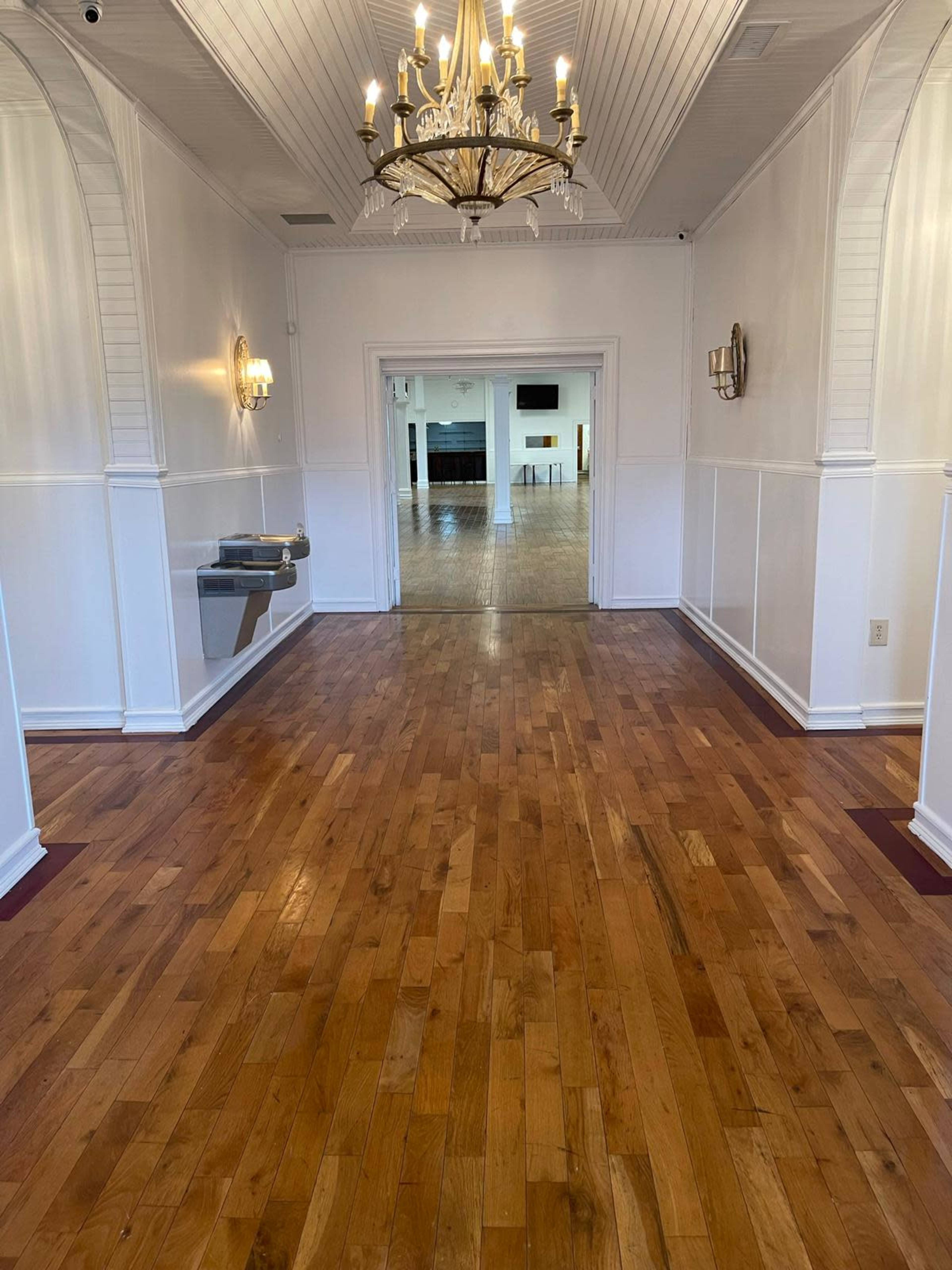 A well-lit hallway with polished wooden floors leads to an open room at the far end, featuring a chandelier and wall sconces.