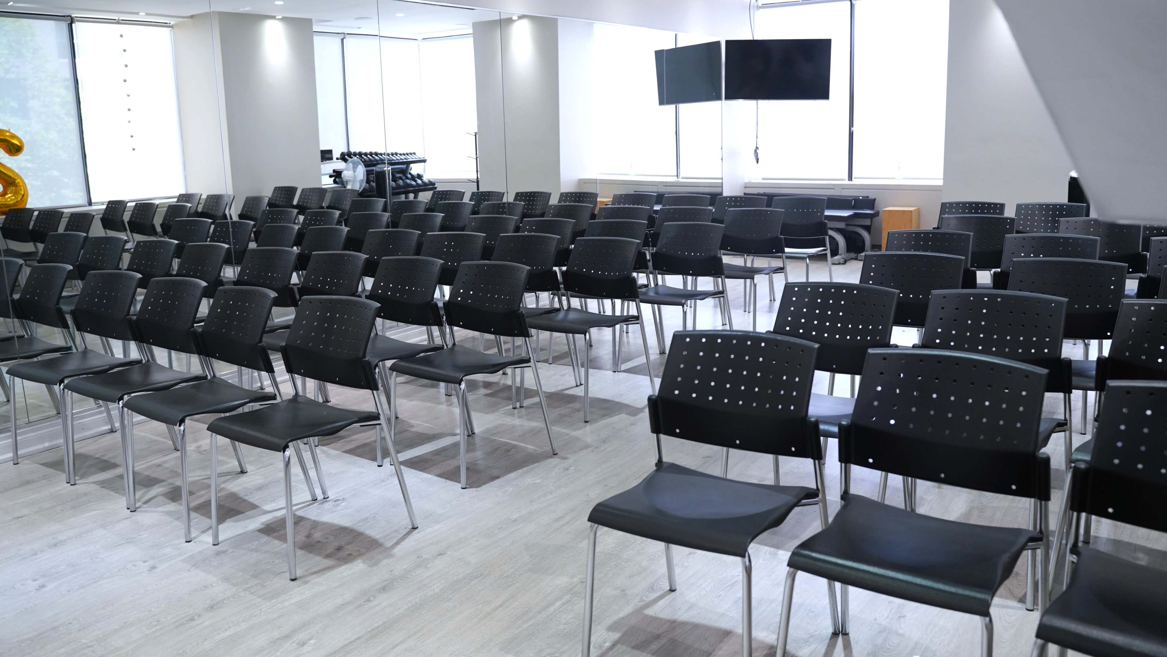 The image shows a spacious room arranged with rows of black, stackable chairs facing a blank television screen and large windows.