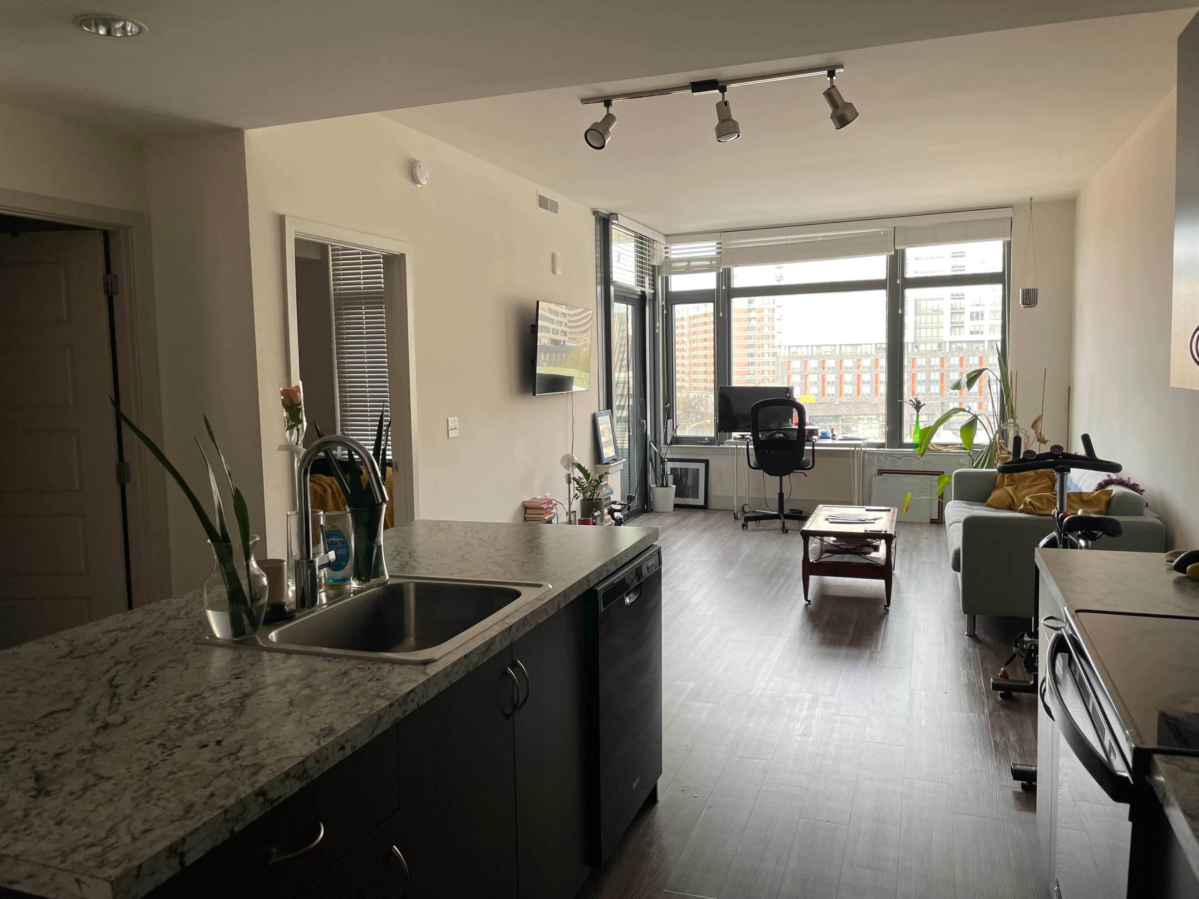 An open-concept apartment interior with a kitchen on the left, a living area with a sofa on the right, and large windows overlooking a cityscape.