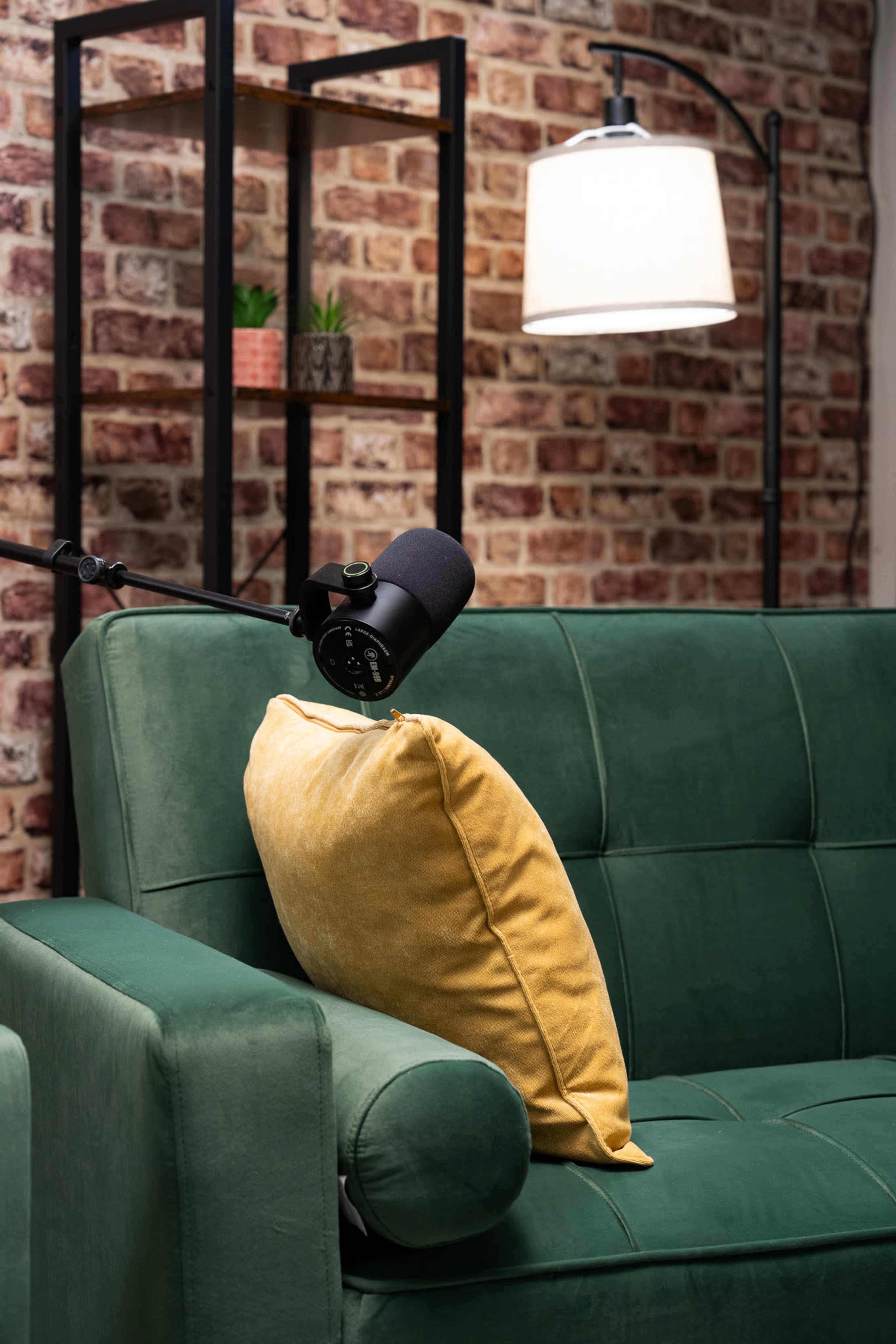 A green sofa with a yellow pillow is positioned in front of a brick wall and a lamp on a nearby shelf.