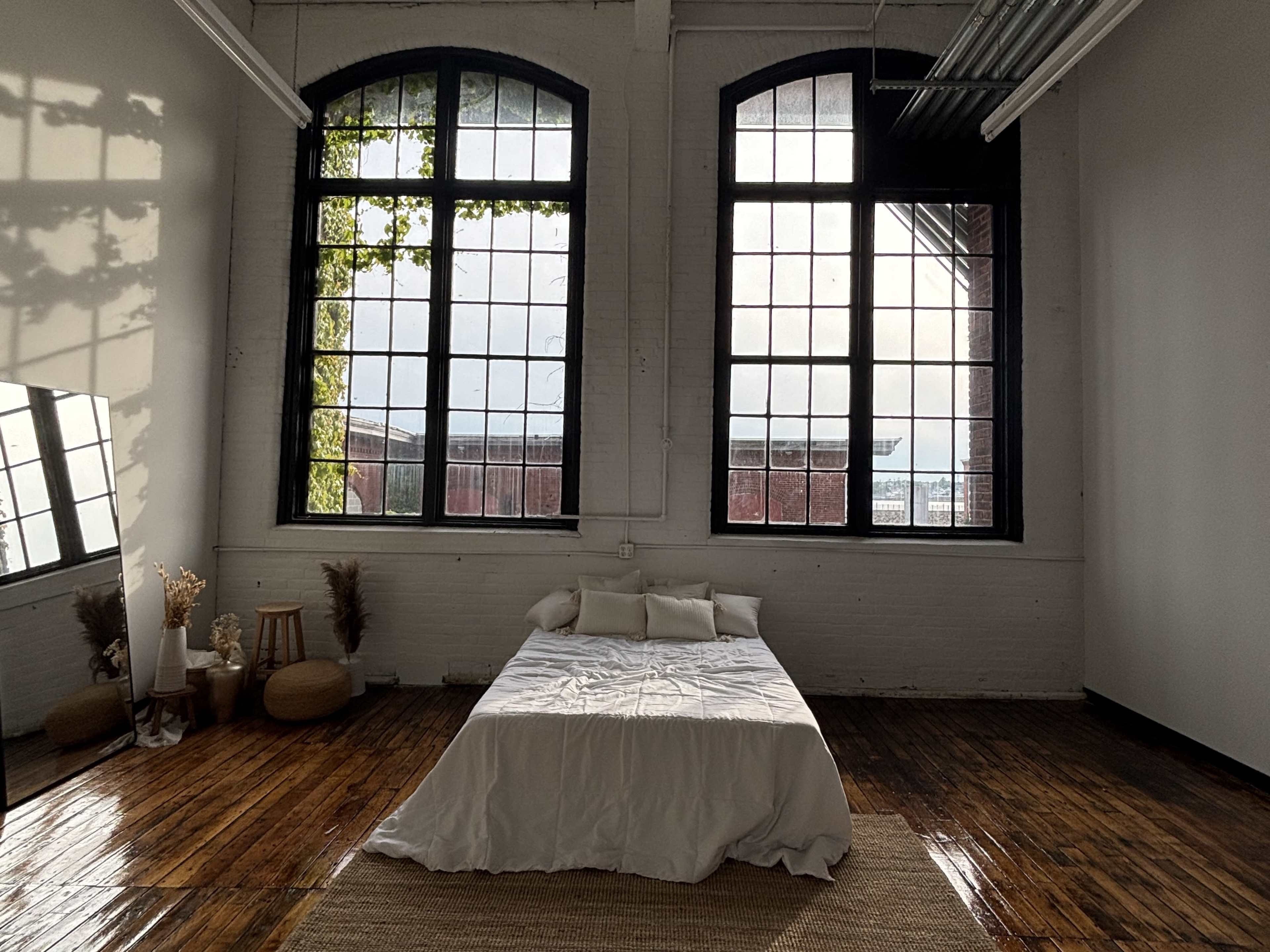 The image shows a spacious room with two large windows, a bed adorned with white linens, and wooden flooring reflecting natural light.