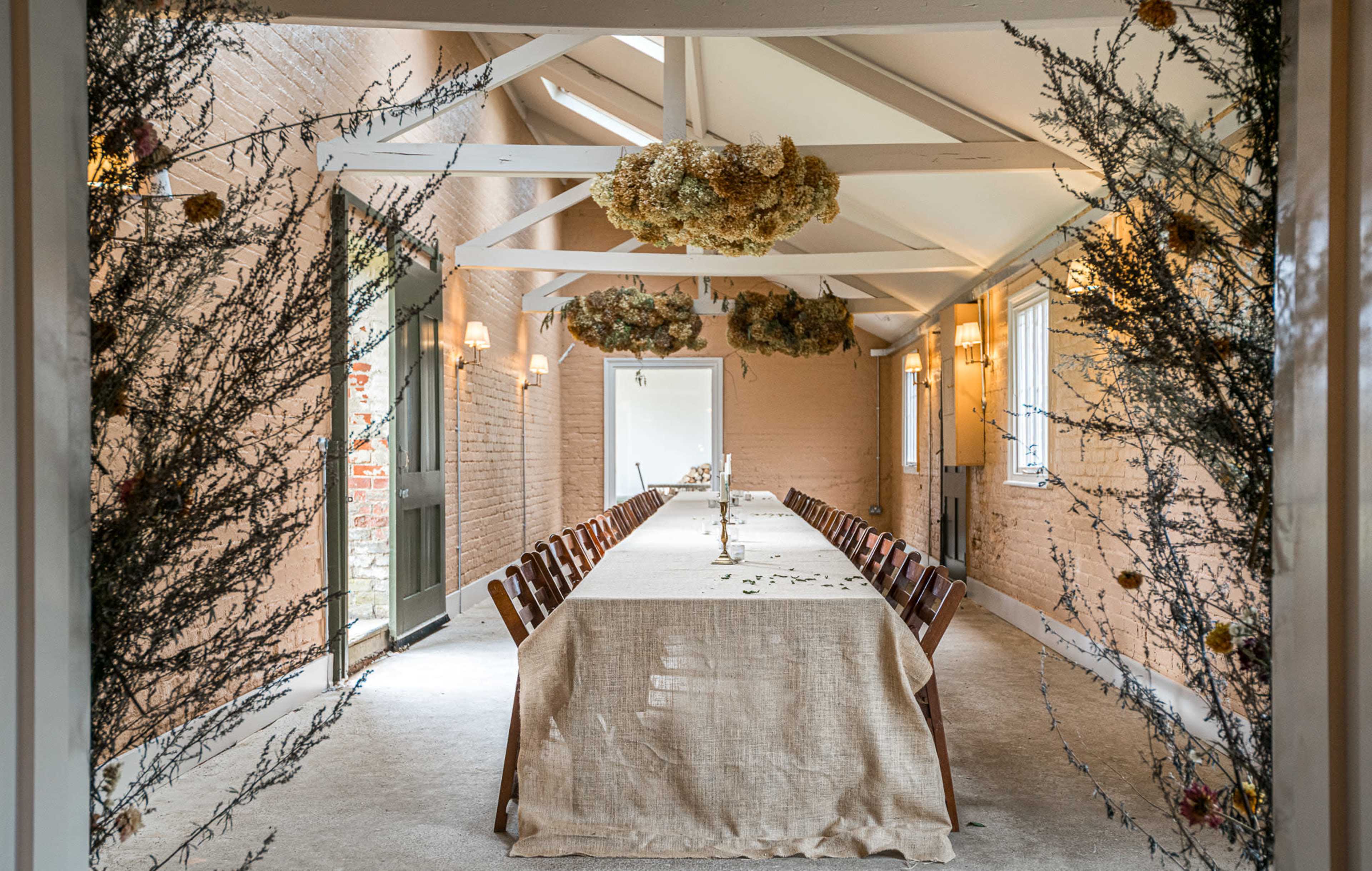A long dining table is set in a rustic room, adorned with dried floral decorations and natural lighting from large windows.
