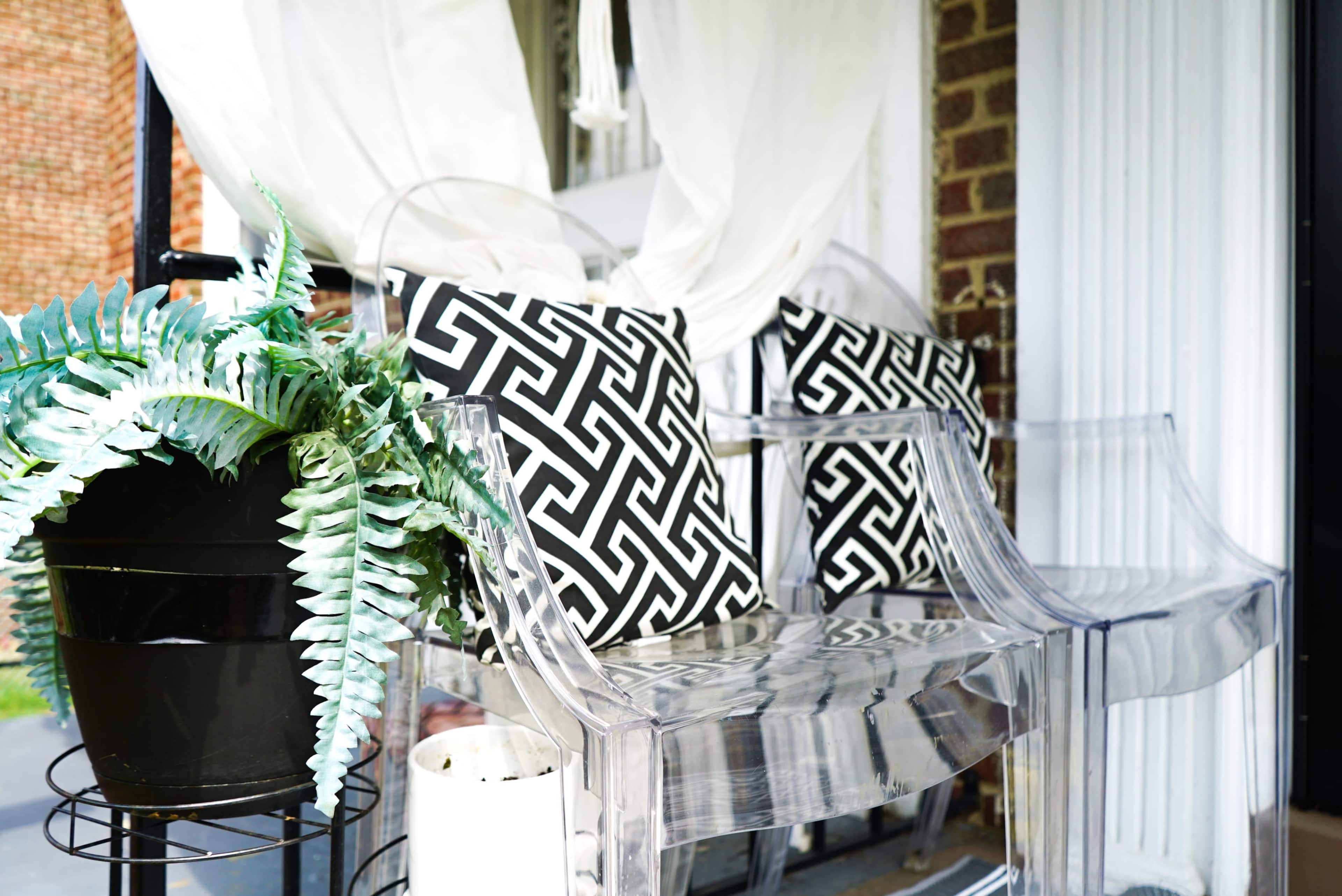 A transparent bench with black and white geometric patterned cushions is placed next to a potted plant on a porch.