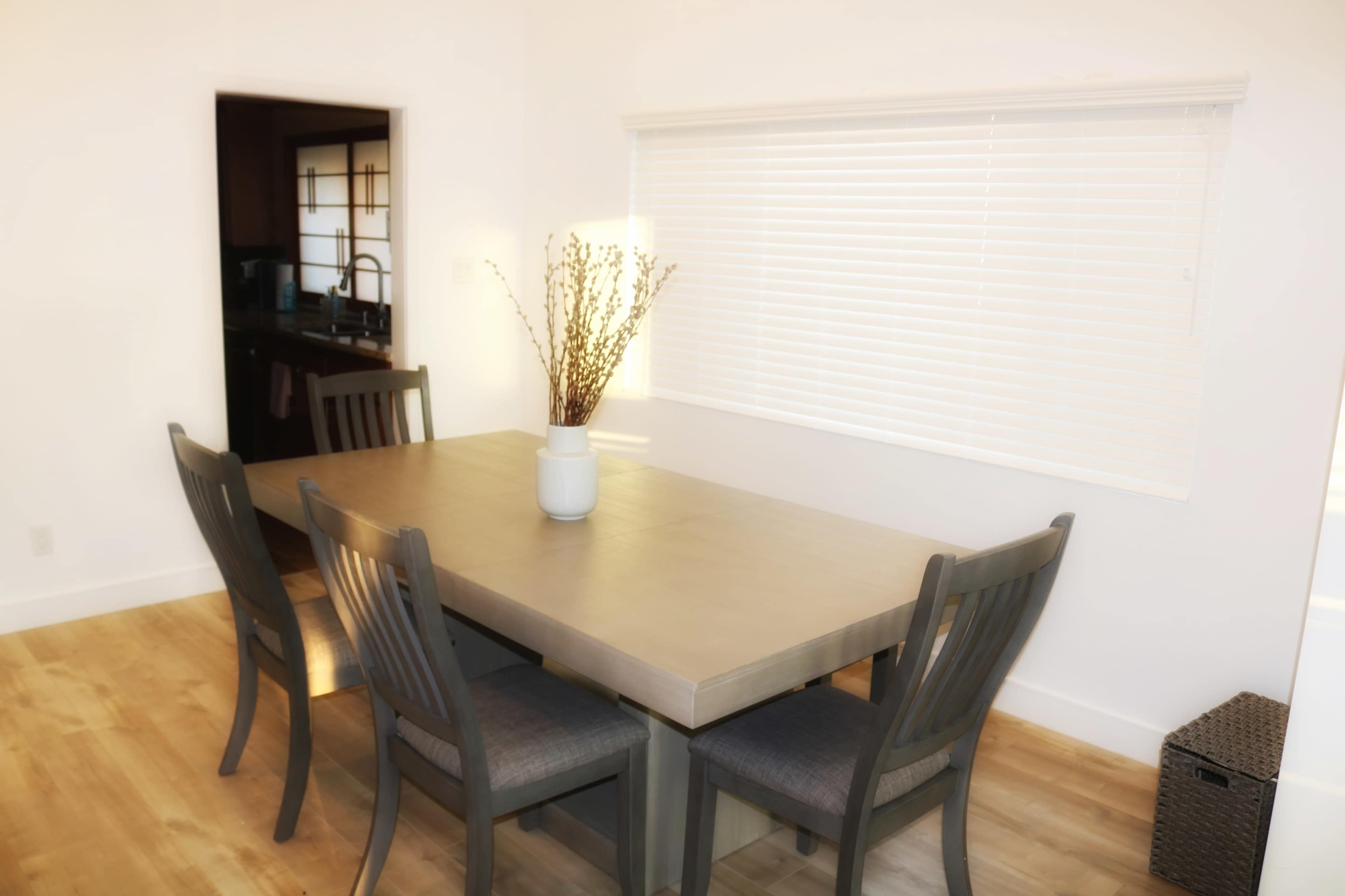A rectangular dining table with four chairs is positioned in a well-lit room featuring a window with blinds and a vase of dried branches in the center.