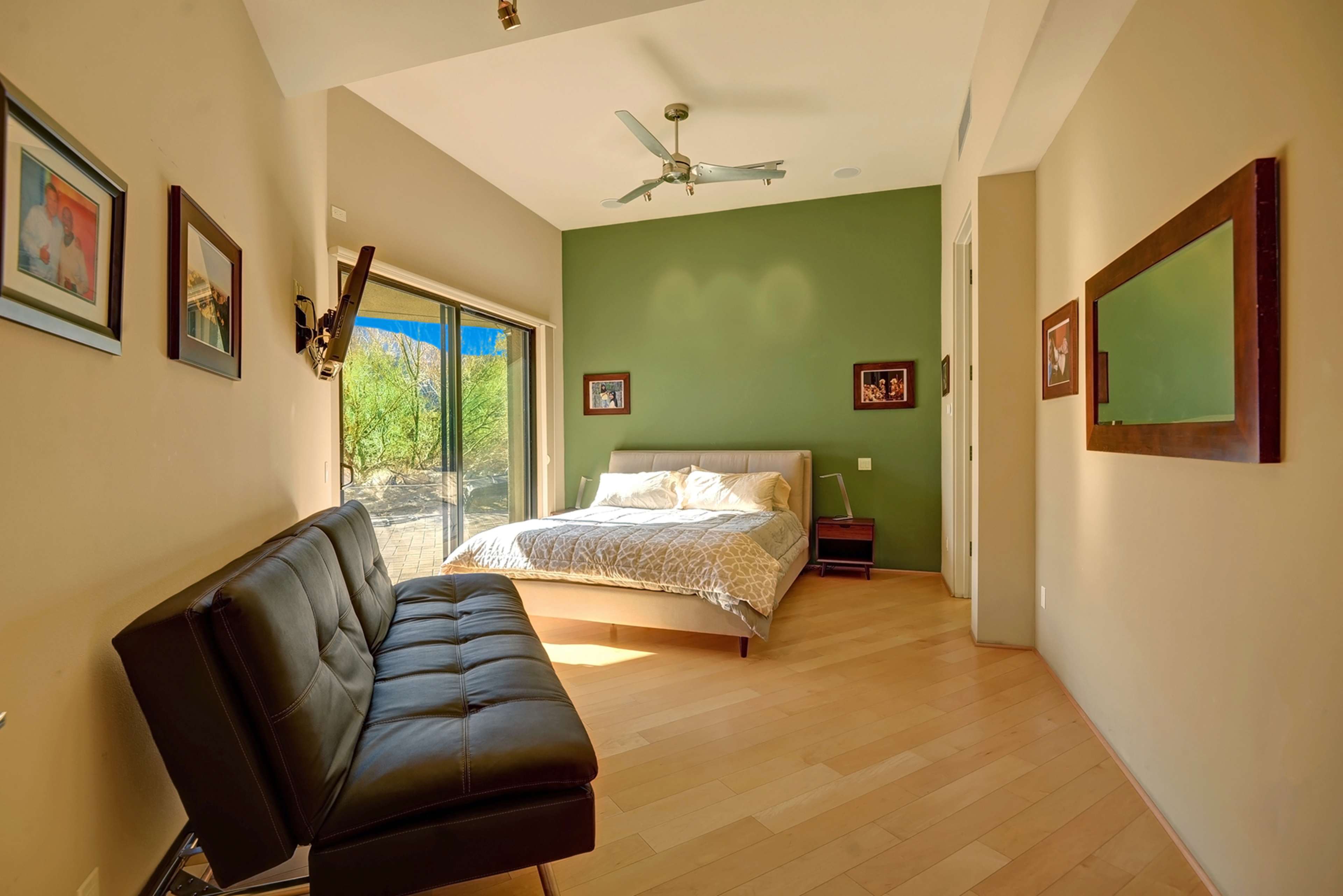 A modern bedroom features a bed with a patterned coverlet, a brown leather sofa, and large windows with views of the outdoors.