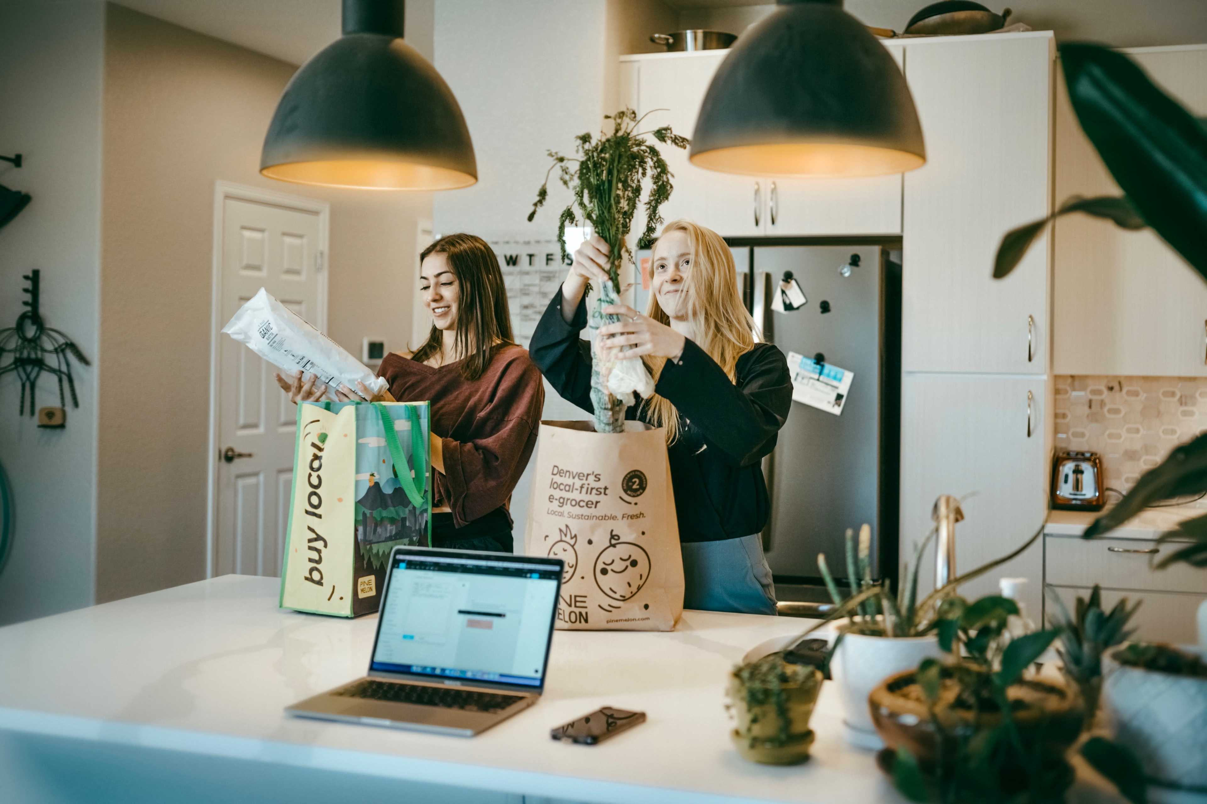 Two women unpack groceries in a modern kitchen while standing at a white countertop.