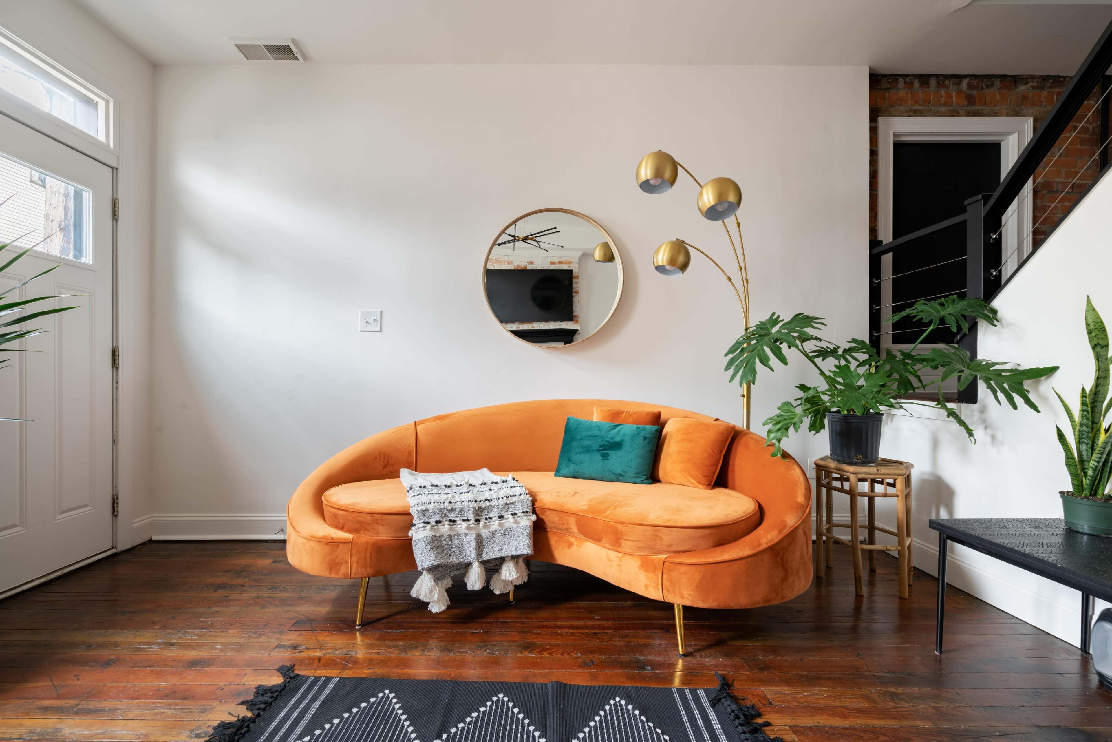 A modern orange curved sofa sits against a white wall, accompanied by a circular mirror, a standing lamp, and various indoor plants in a well-lit living space.